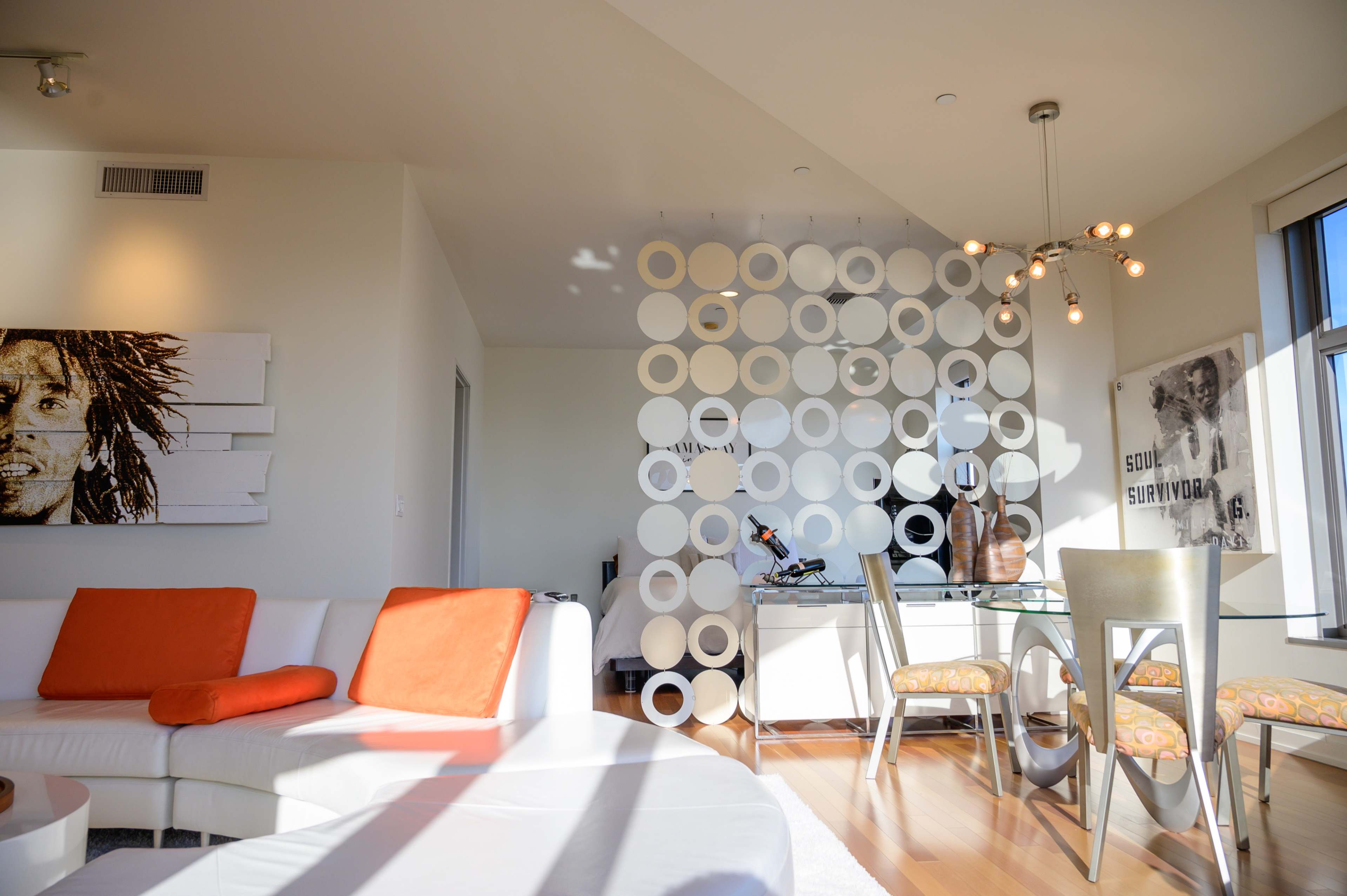 A modern living room with a circular decorative wall, a white sectional sofa with orange pillows, a dining area featuring glass-top tables and metal chairs, and musical elements in the decor.