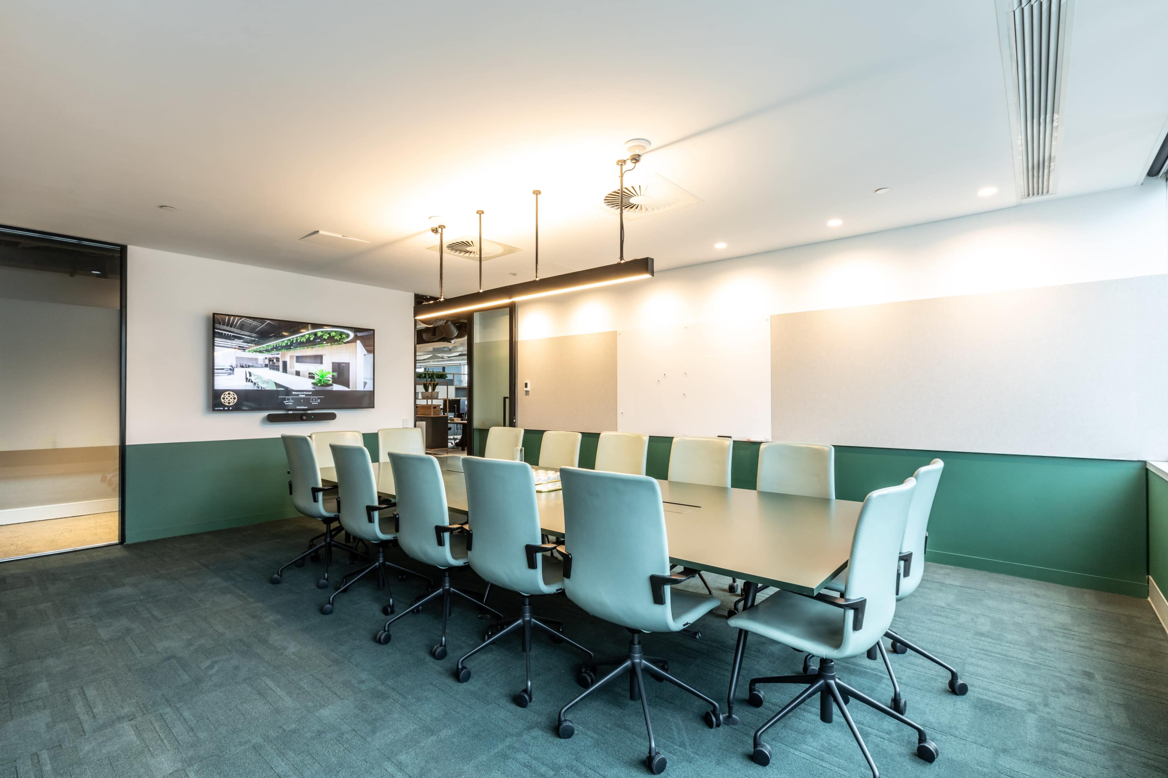 A conference room features a long table surrounded by green chairs and a large wall-mounted screen.