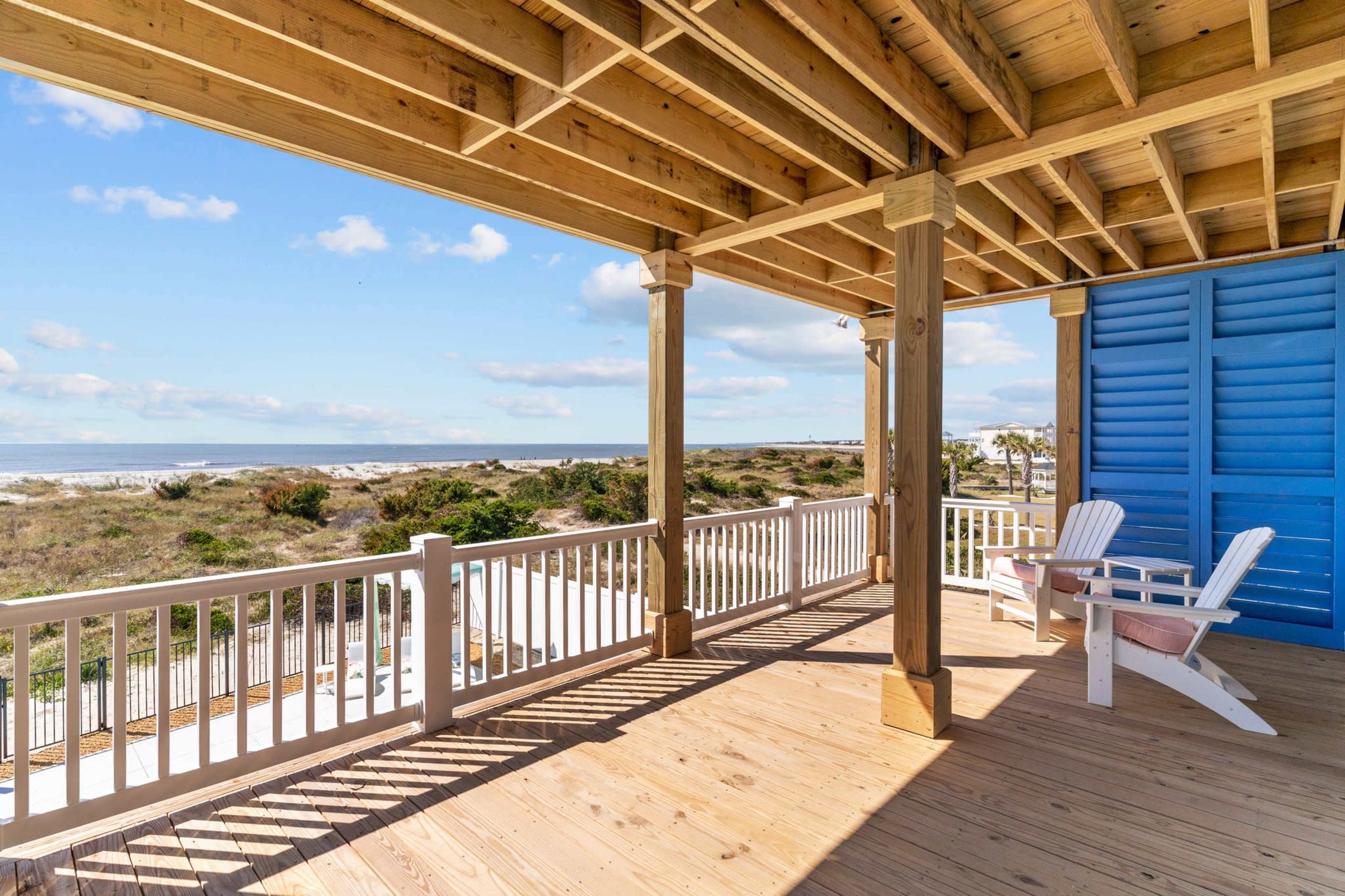 A wooden deck with white railings and two white chairs, overlooking a beach with dunes and a clear sky.