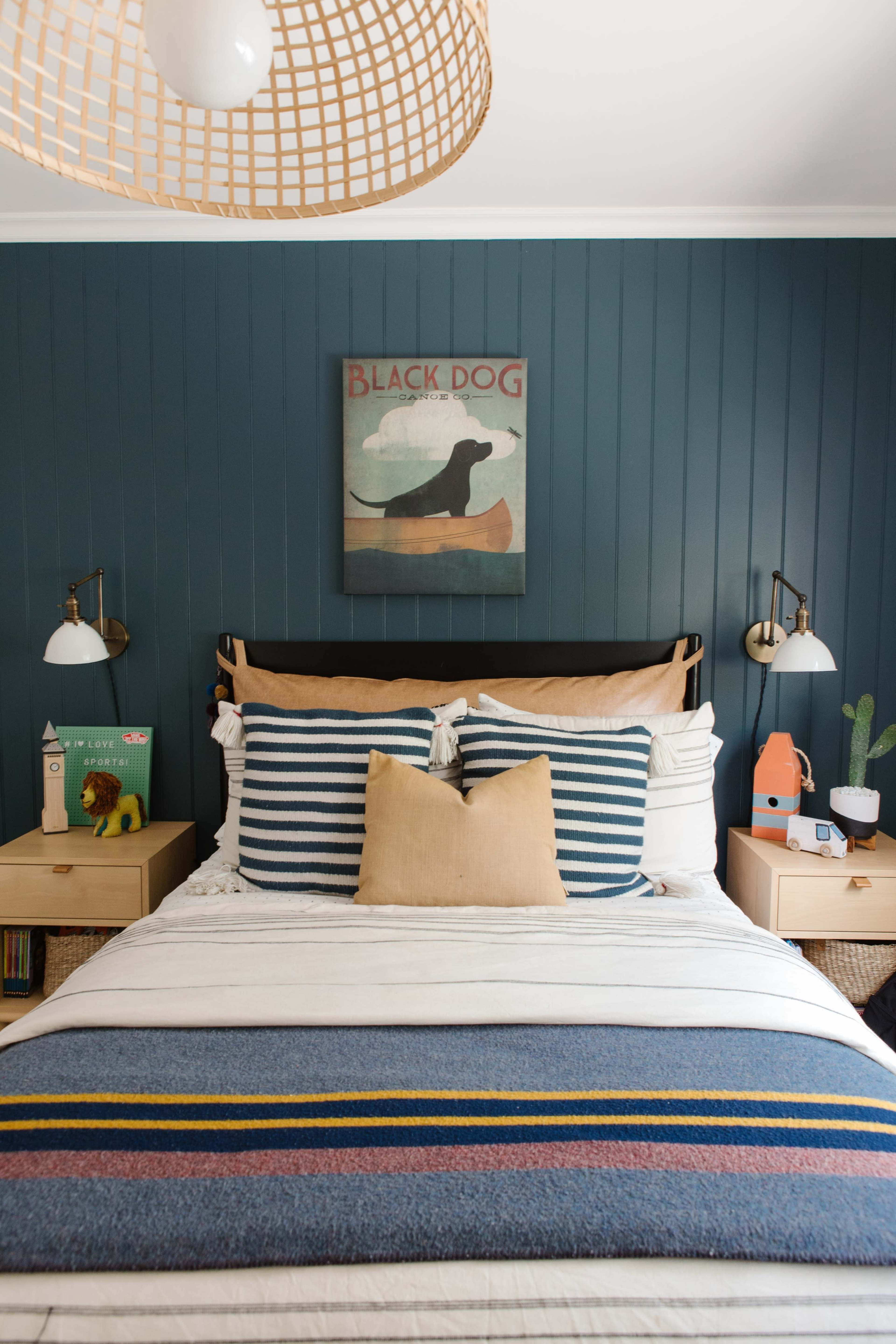 A neatly arranged bedroom featuring a bed with striped pillows and a woven pendant light above, alongside nightstands with decorative items and a dark blue accent wall.