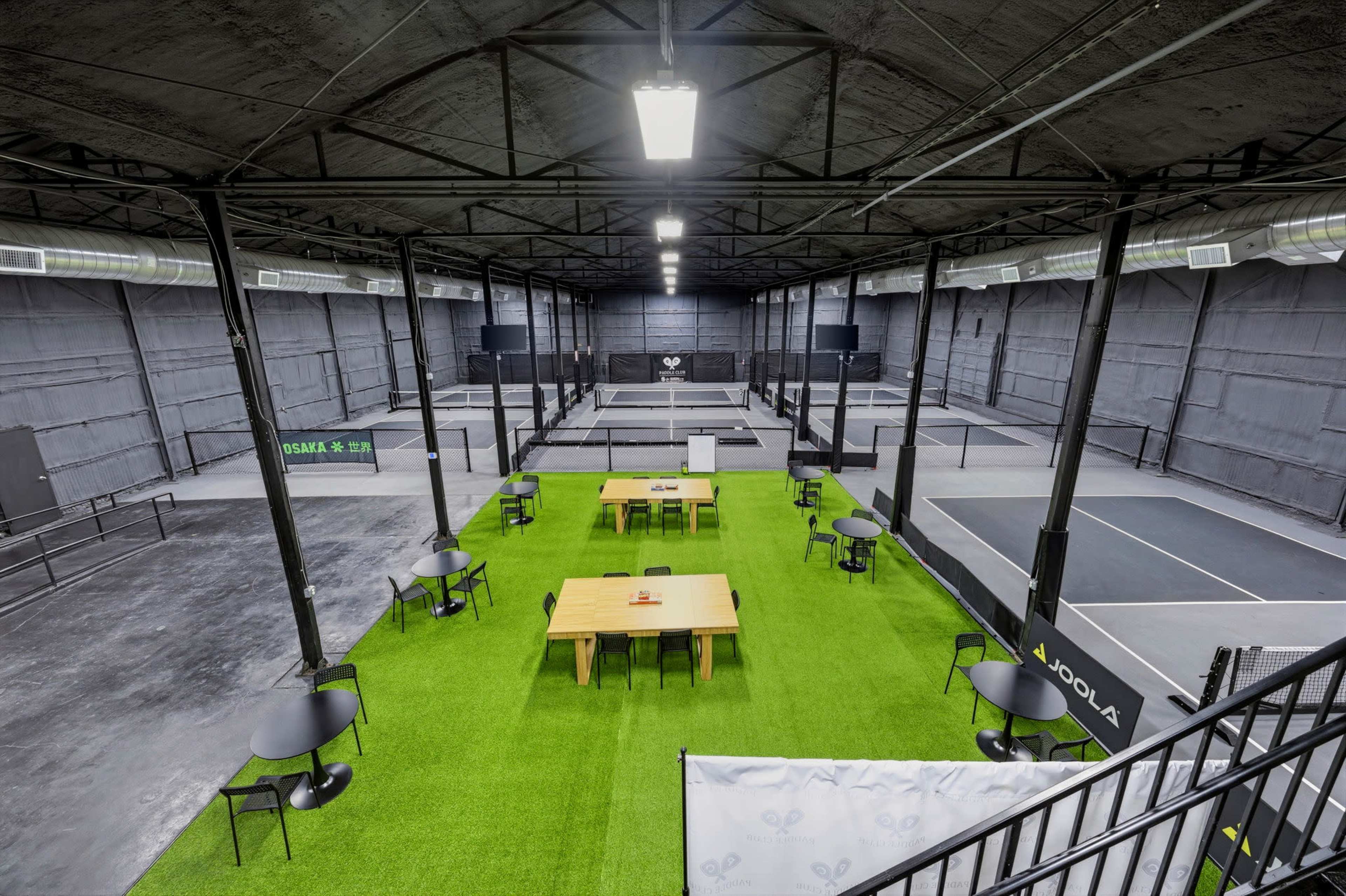 The image shows a spacious indoor sports facility featuring multiple pickleball courts surrounded by seating areas with tables and chairs on green turf.