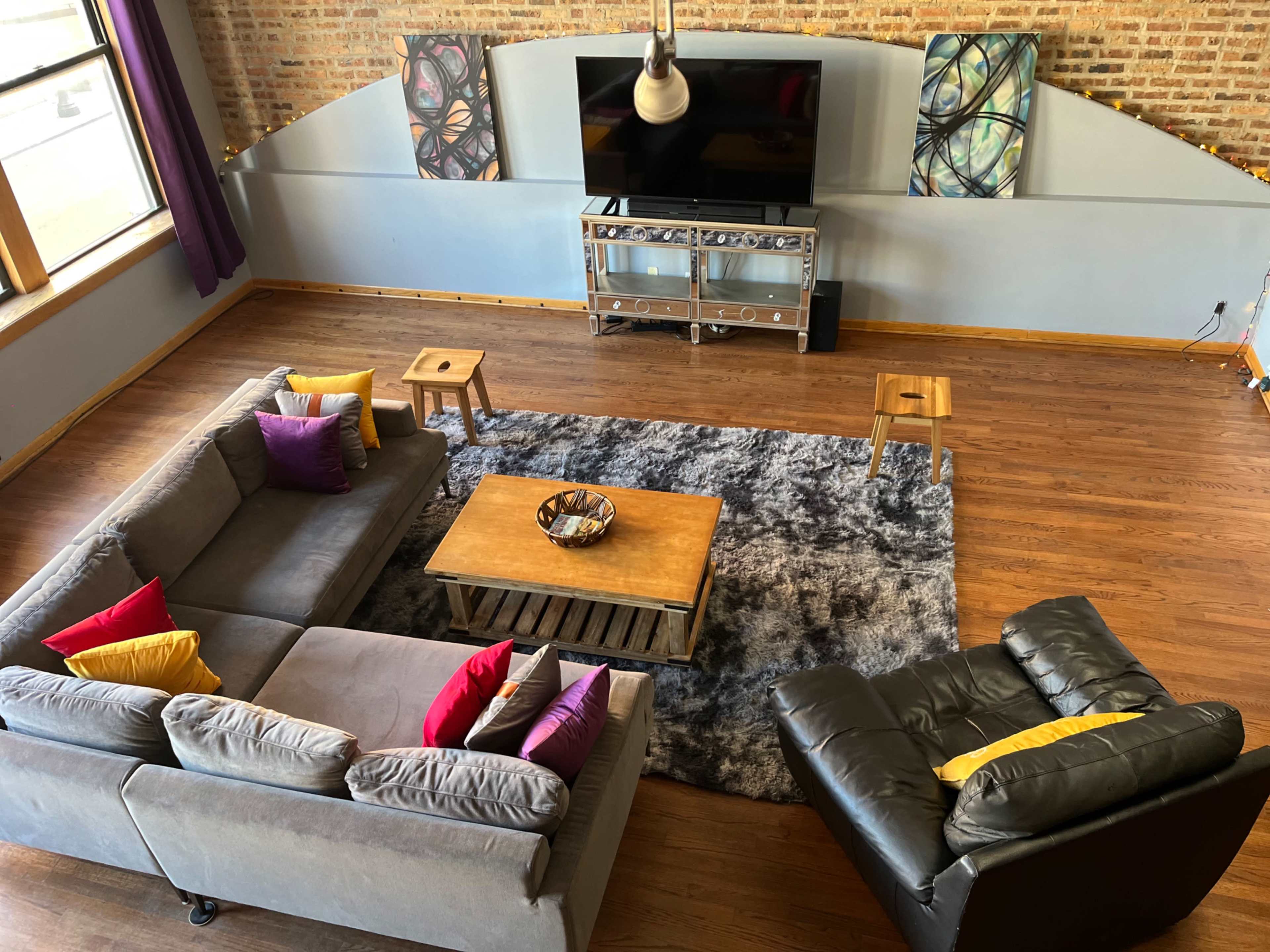 The image shows a spacious living room with a gray sectional sofa, a black armchair, a wooden coffee table on a plush rug, and a television mounted on the wall above a modern console table.