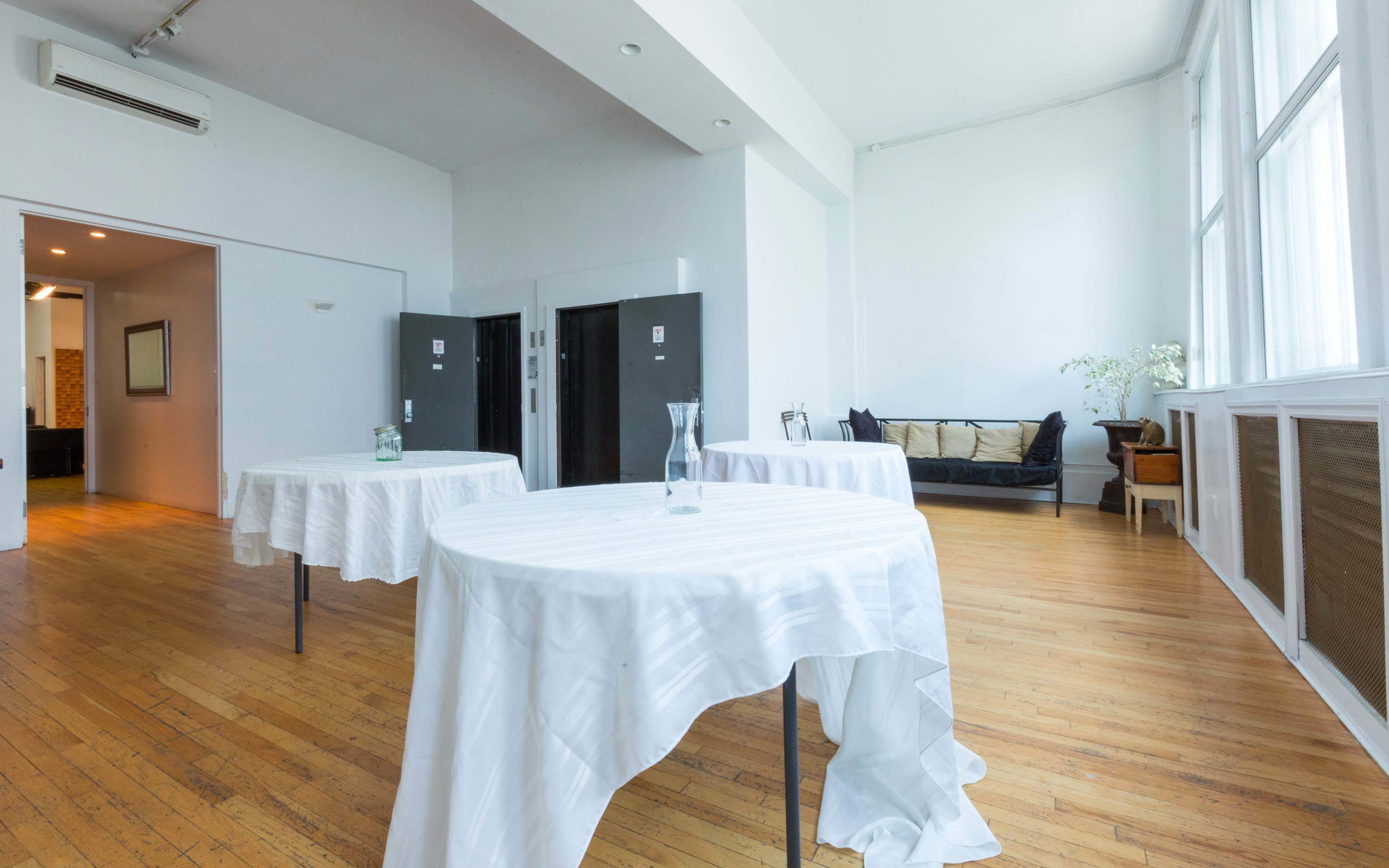 The image shows a spacious room with wooden floors, several round tables covered in white tablecloths, and large windows allowing natural light to enter.