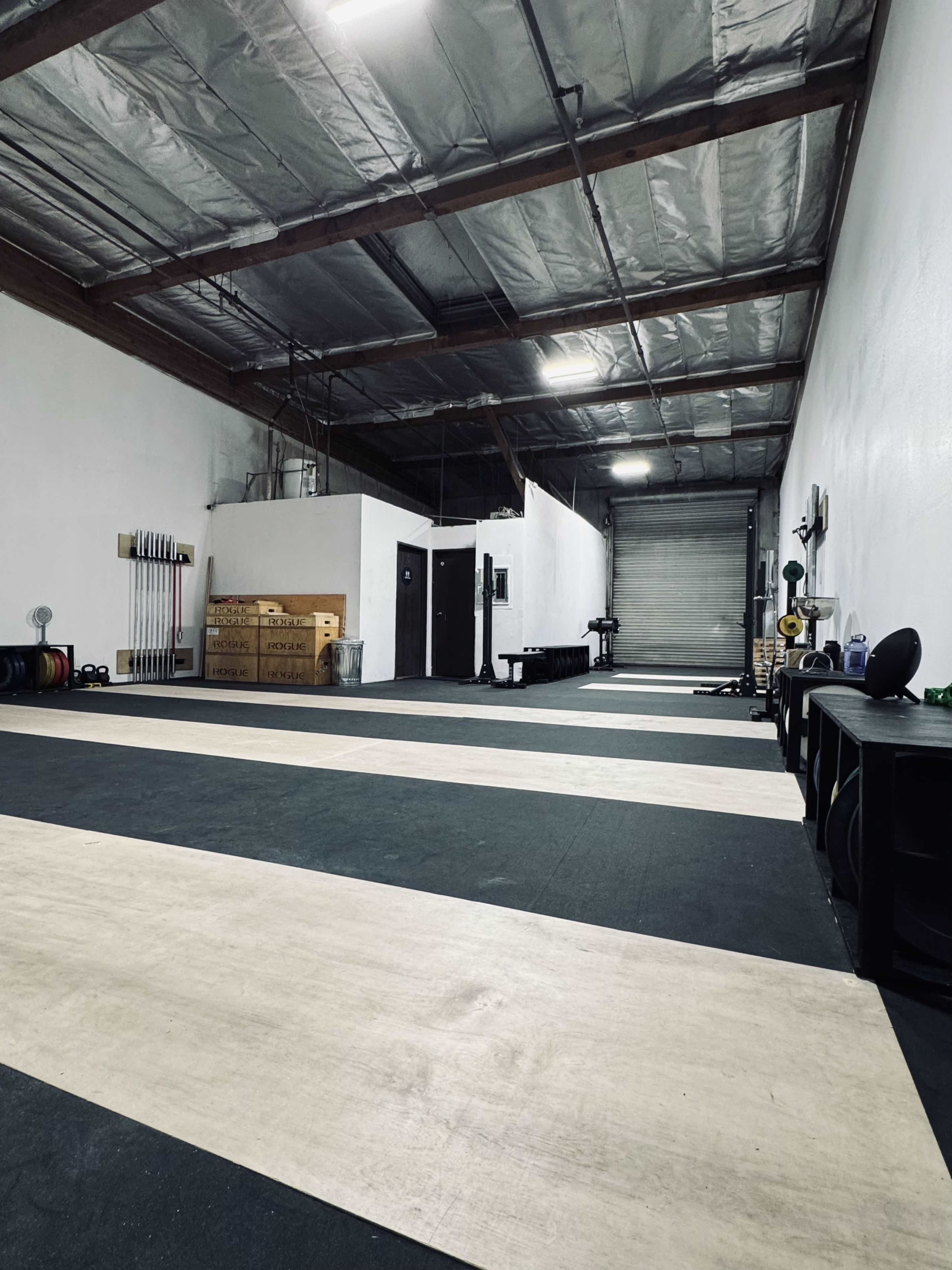 The image shows a spacious indoor gym with a marked exercise area, weightlifting equipment, and a loading dock at the far end.