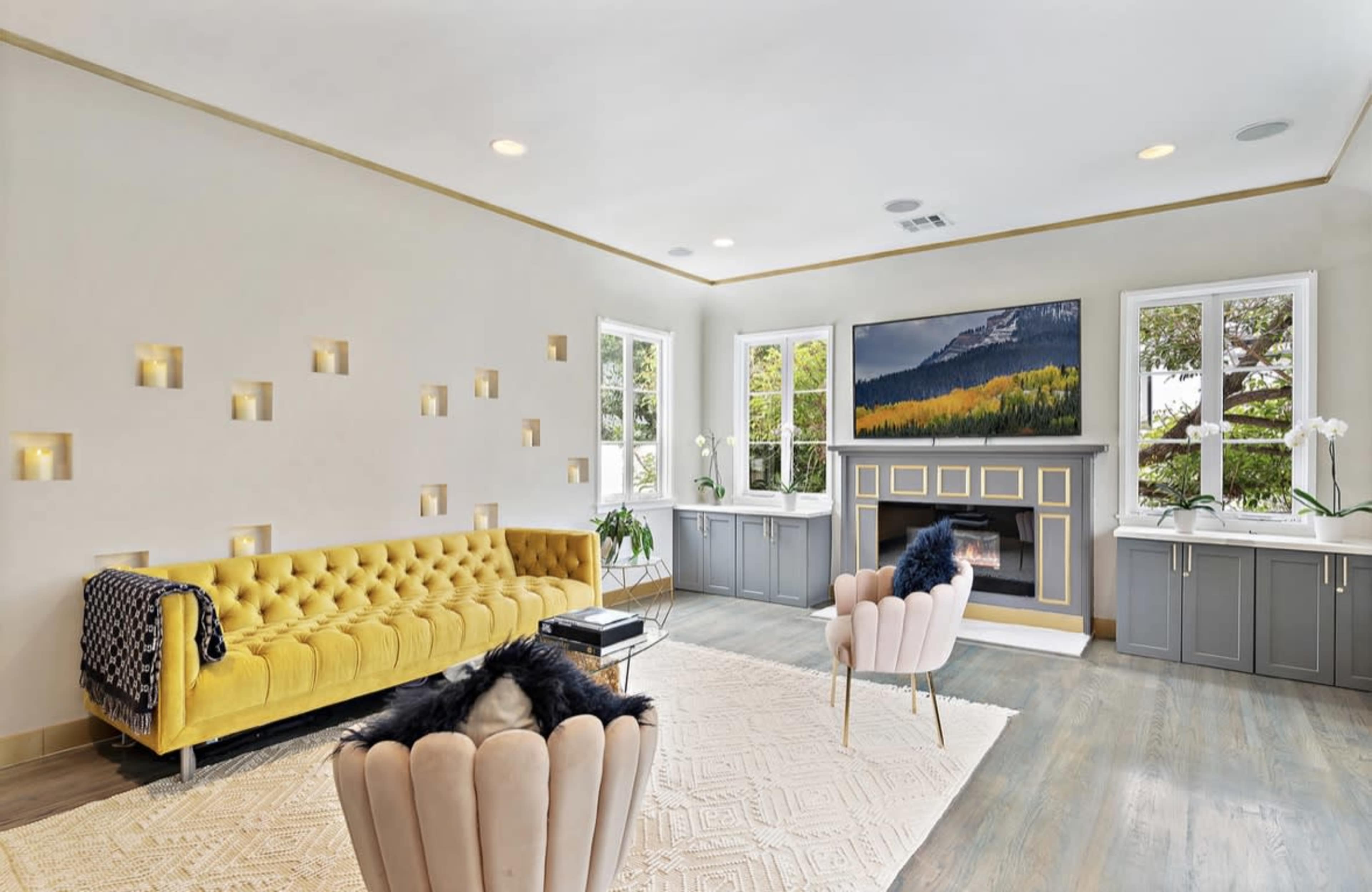 The living room features a yellow tufted sofa, two pink velvet chairs, a wall with illuminated niches, and large windows overlooking greenery.