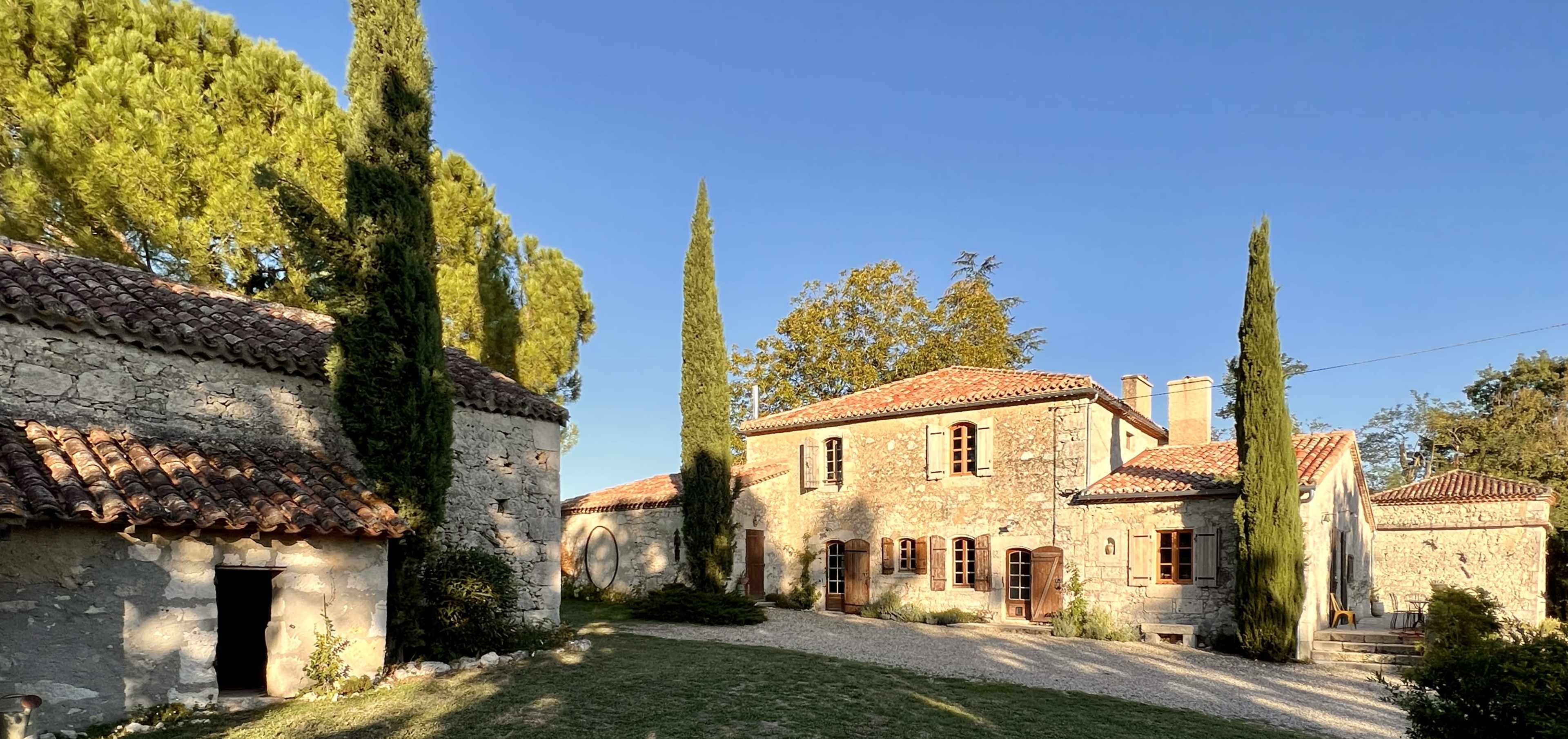 A stone house with a tiled roof surrounded by tall cypress trees and a gravel pathway.