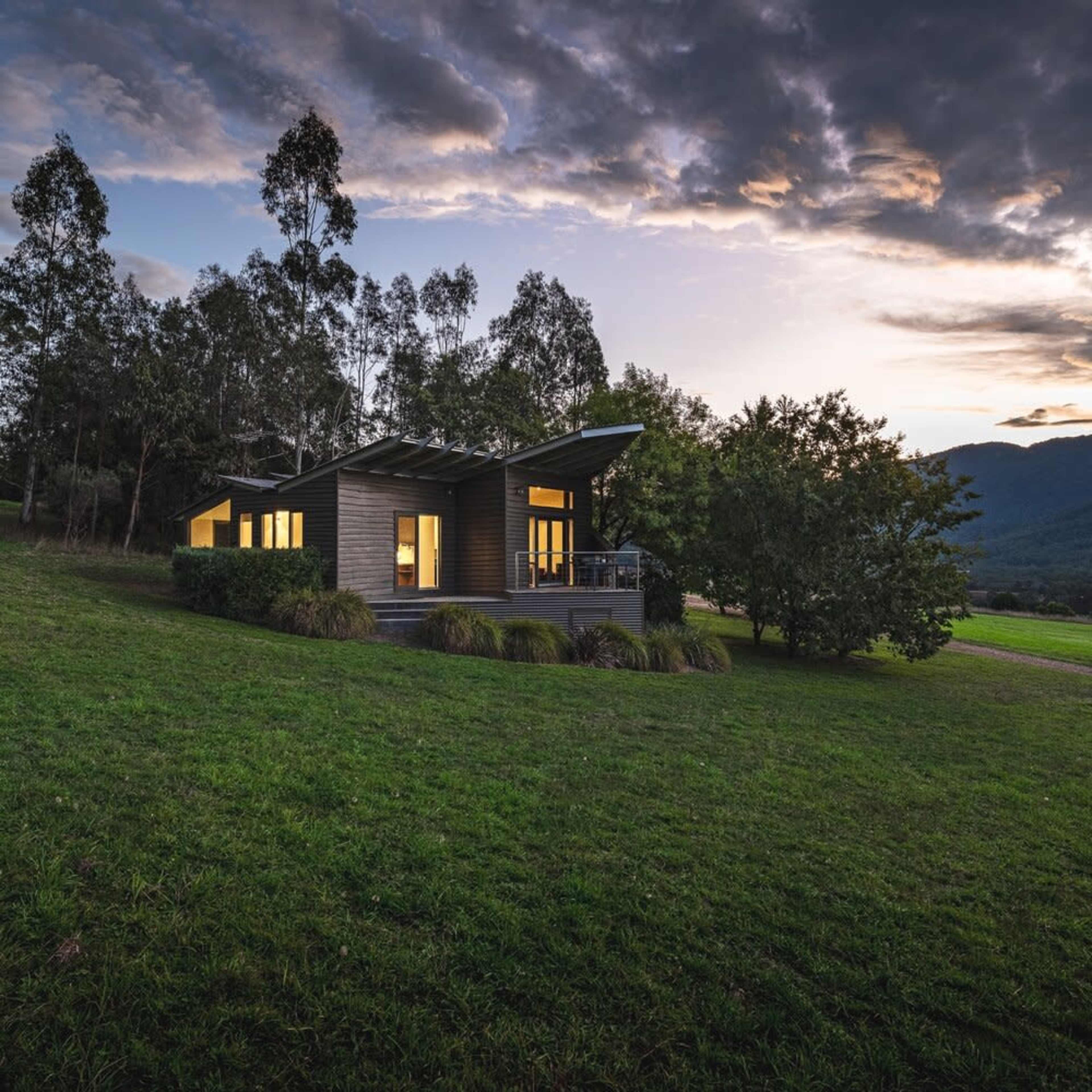 A modern wooden house with large windows and a sloped roof is situated on a grassy hillside surrounded by trees at sunset.