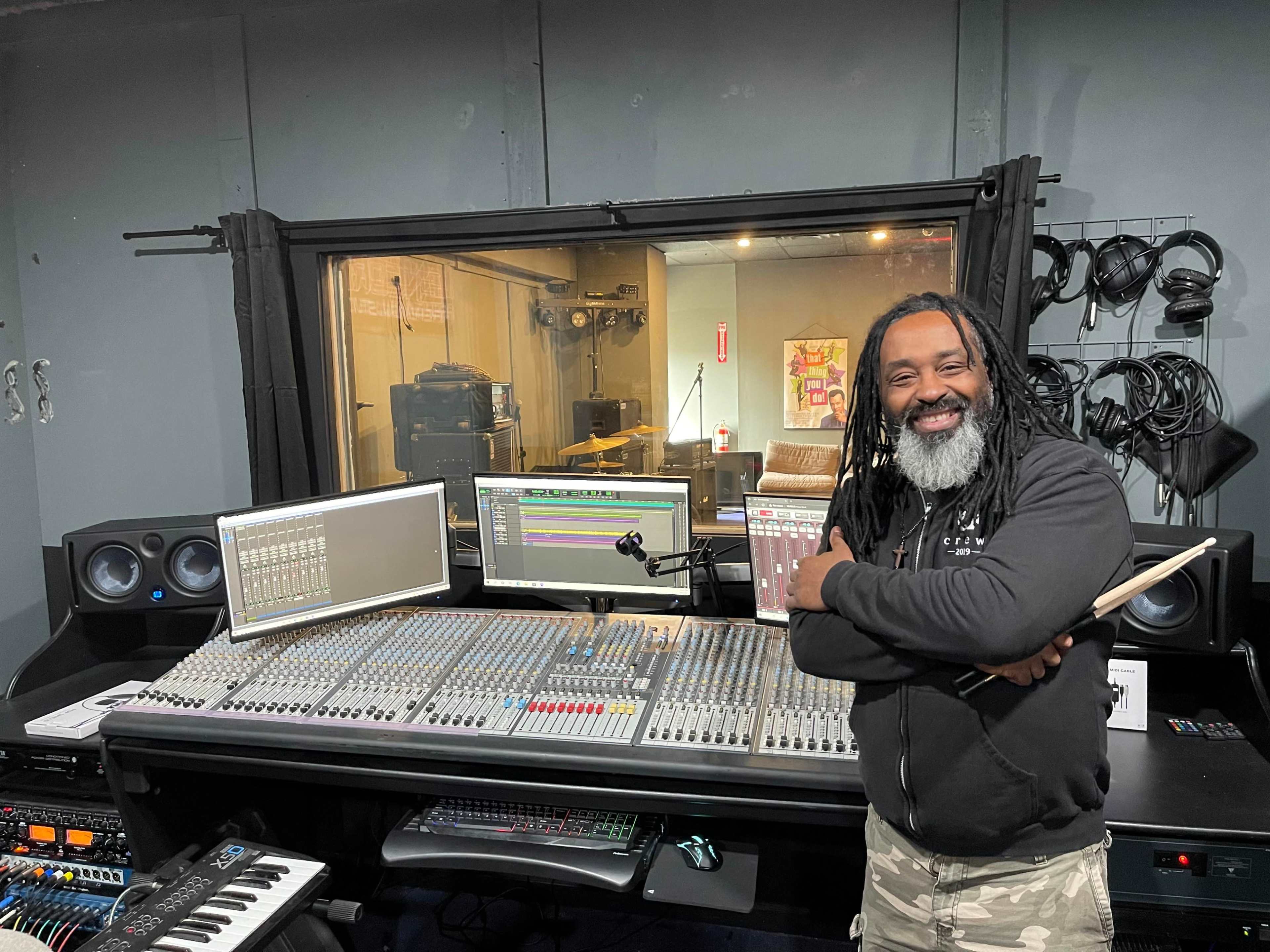 A man stands in front of a large audio mixing console with multiple screens in a recording studio.