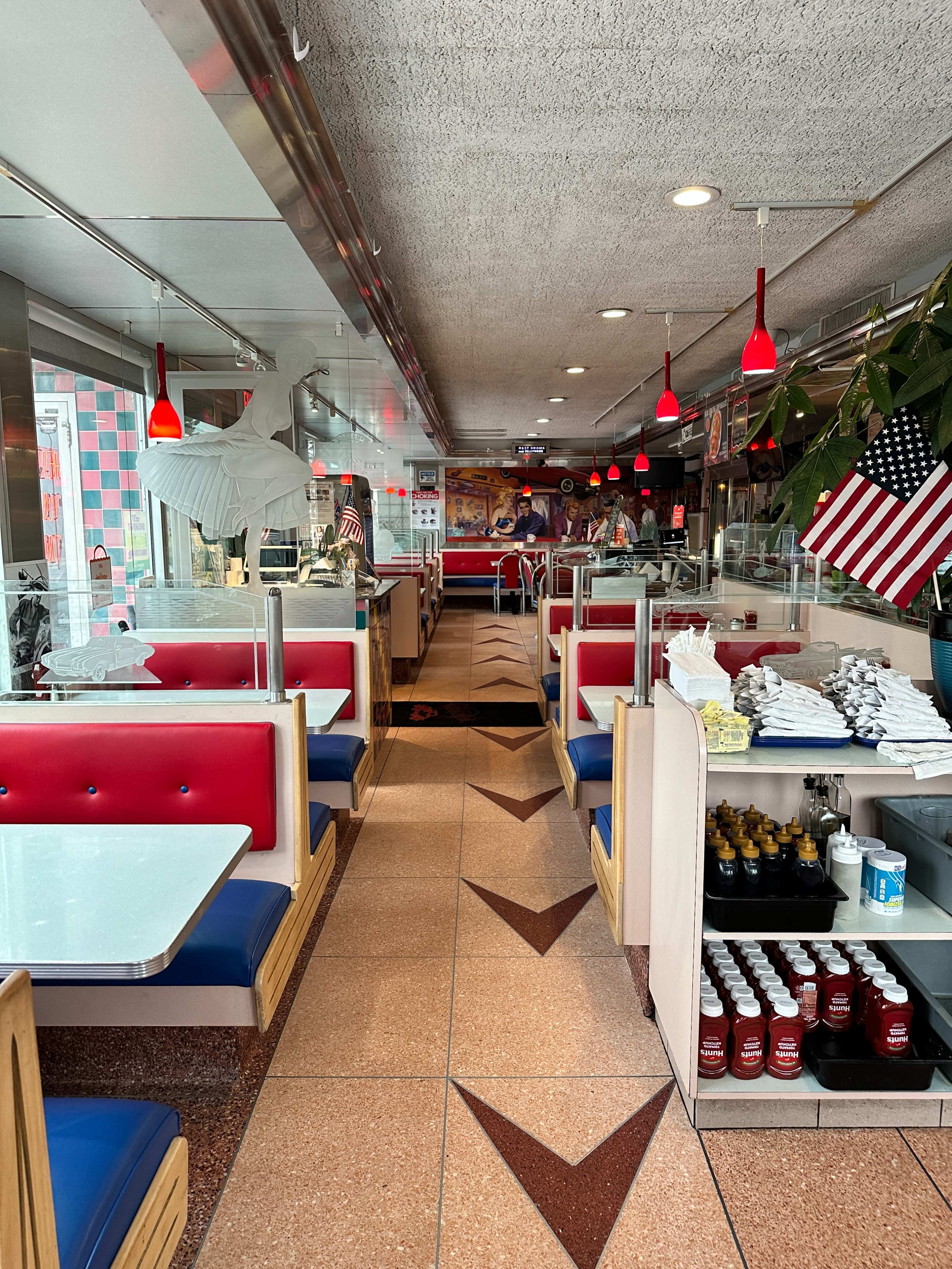 The interior of a diner features red and blue booths, a tiled floor with decorative patterns, and various condiments on display.