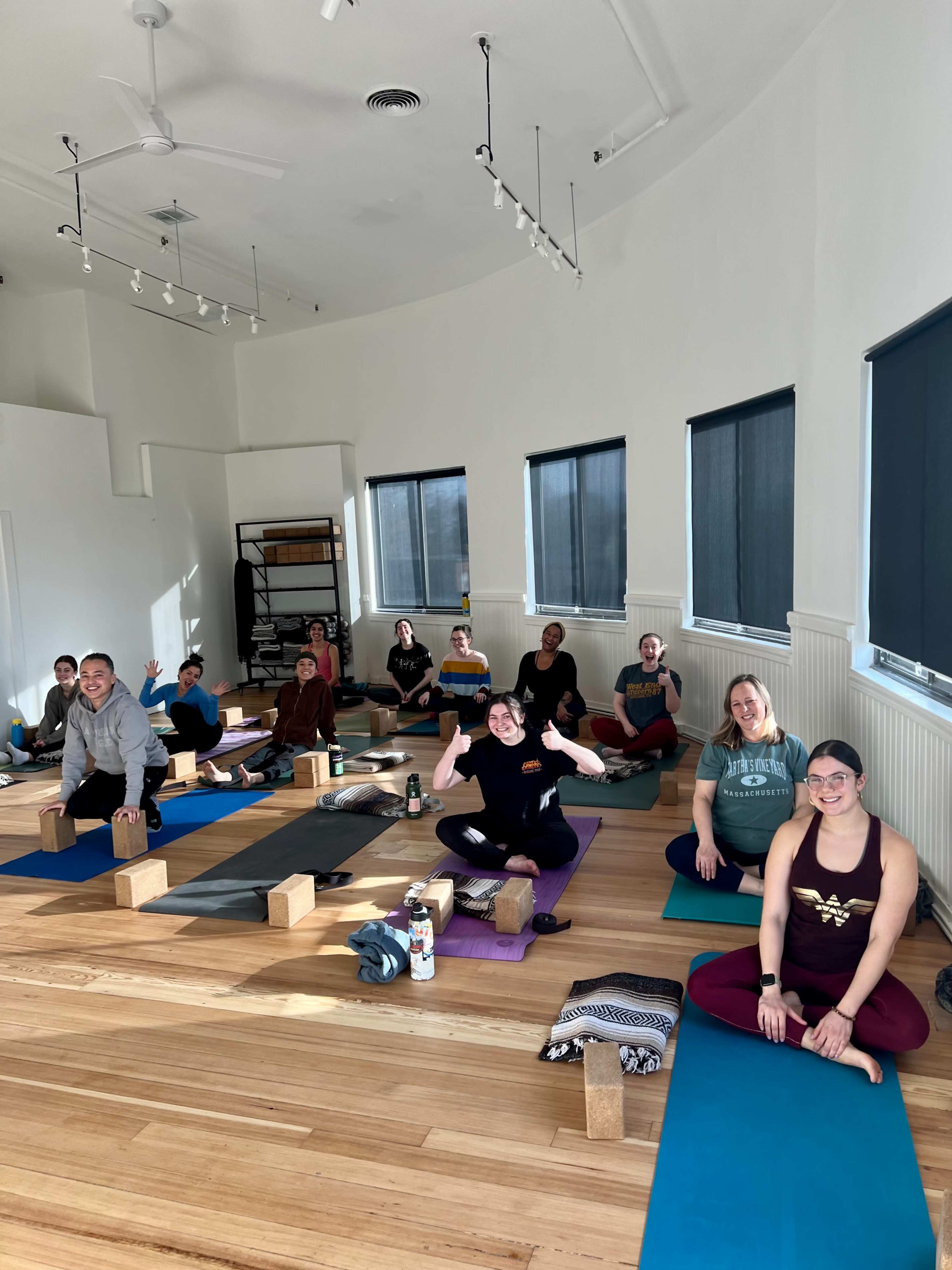 A group of people is sitting on yoga mats in a well-lit studio, with yoga blocks arranged nearby.