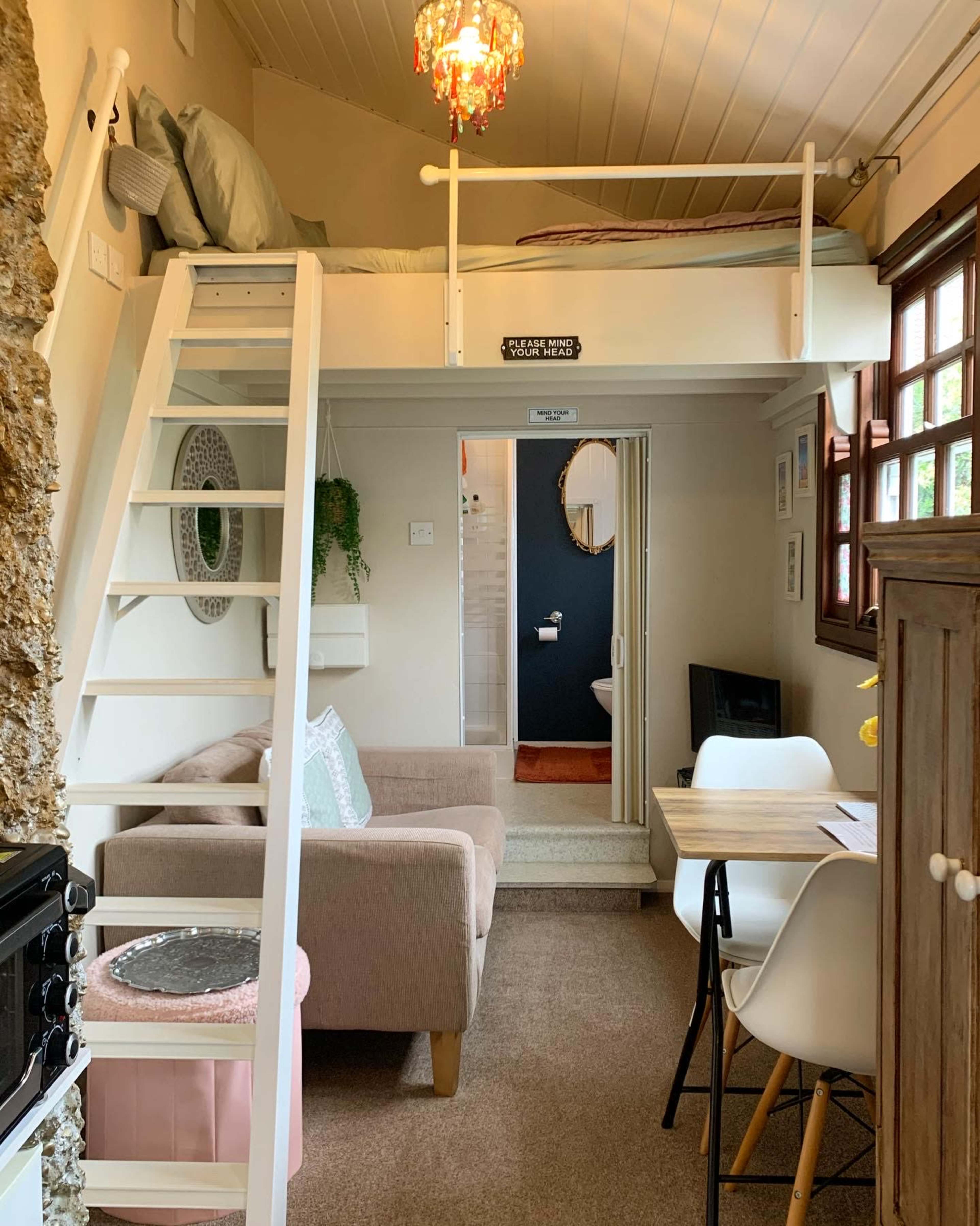 The image depicts a small, cozy interior of a living space featuring a lofted sleeping area, a sofa, a dining table, and a kitchenette.