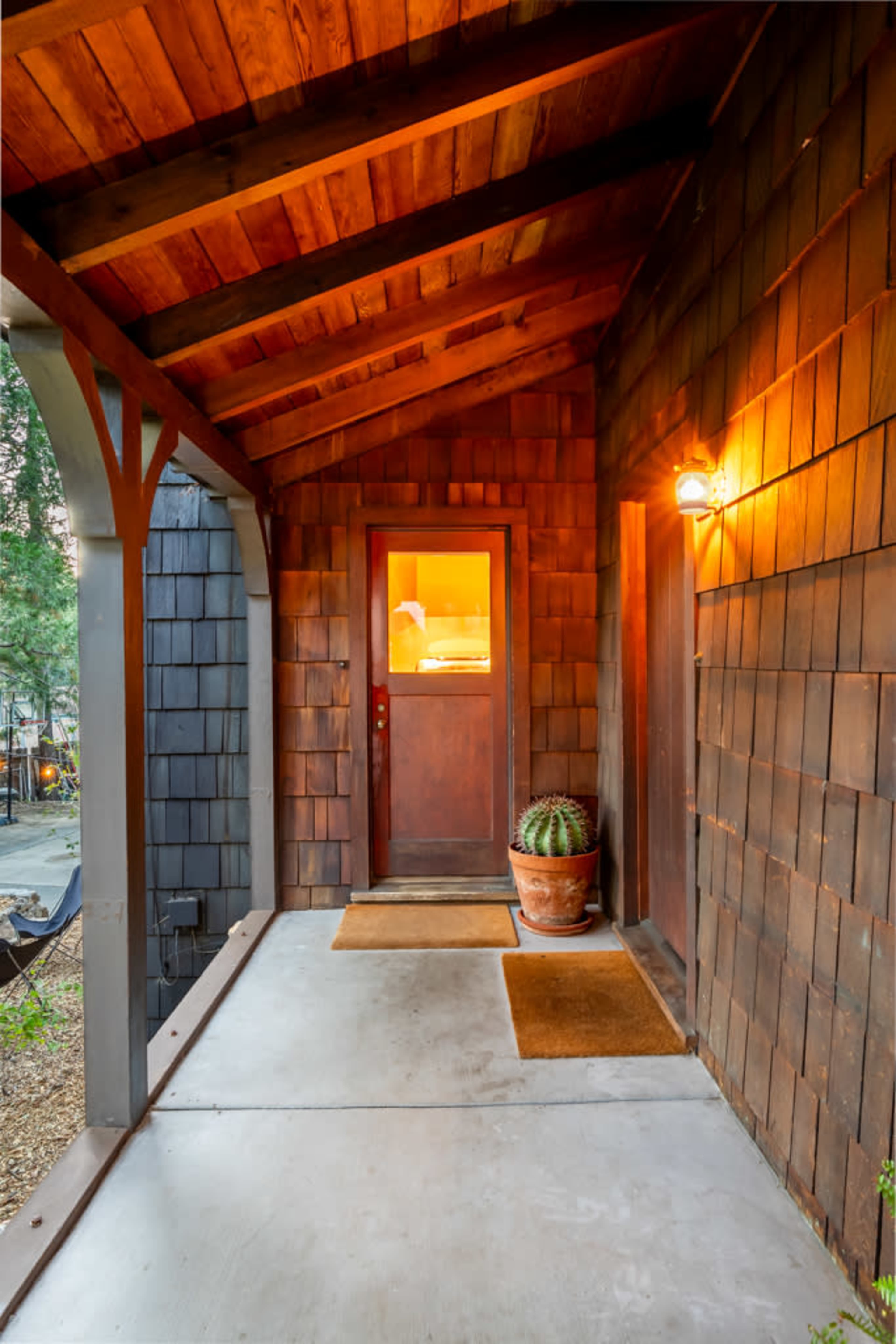 The image shows a wooden porch with a sloped roof, featuring a front door illuminated by warm light and a potted cactus on the side.