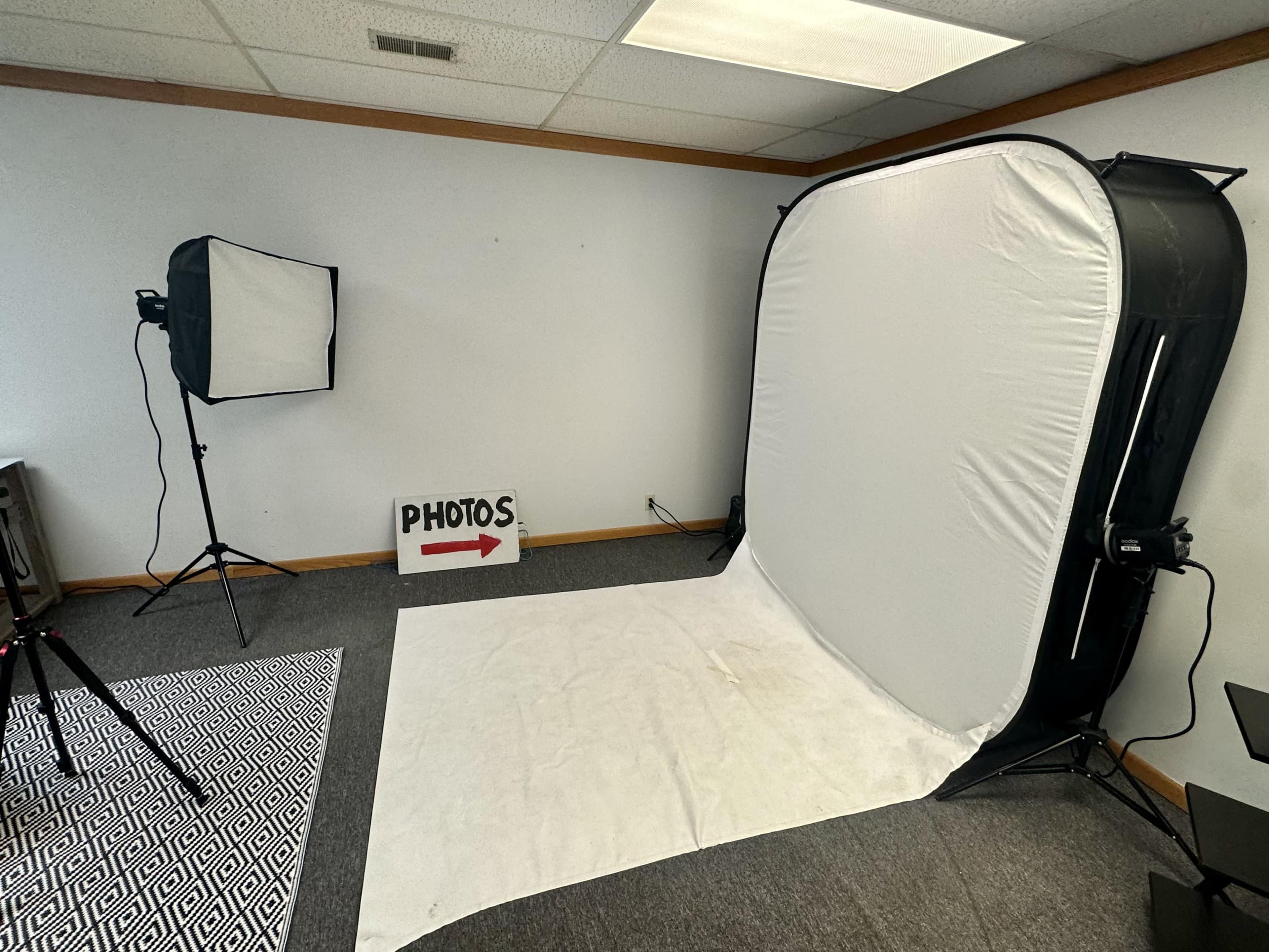 The image shows a photography studio setup featuring a white backdrop, lighting equipment, and a sign indicating the area for photos.
