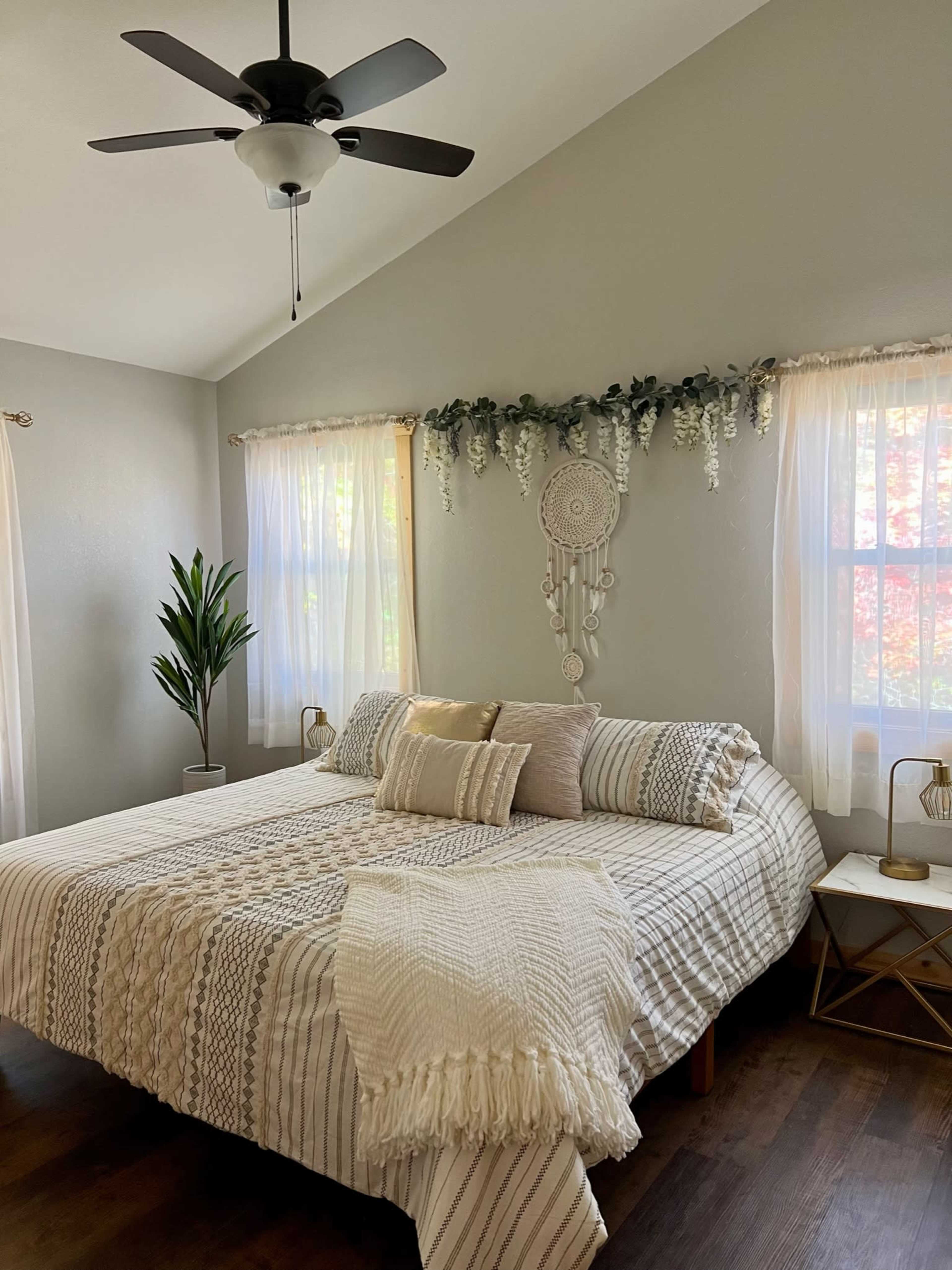 A neatly made bed with patterned bedding is positioned in a well-lit bedroom featuring a ceiling fan and decorative wall hangings.