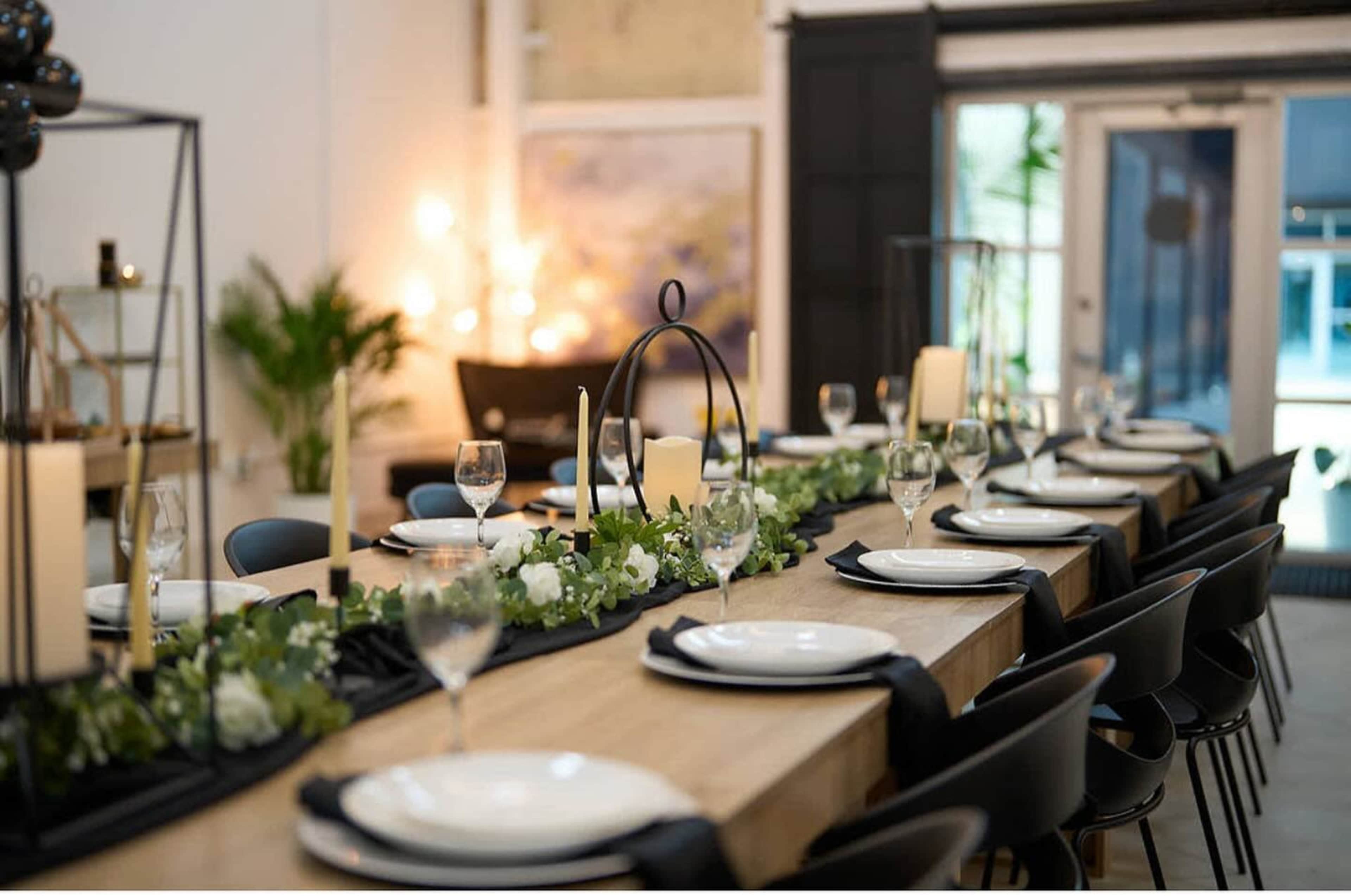 A long dining table is set with white plates, wine glasses, and candles, surrounded by greenery and black napkins in a stylish interior.