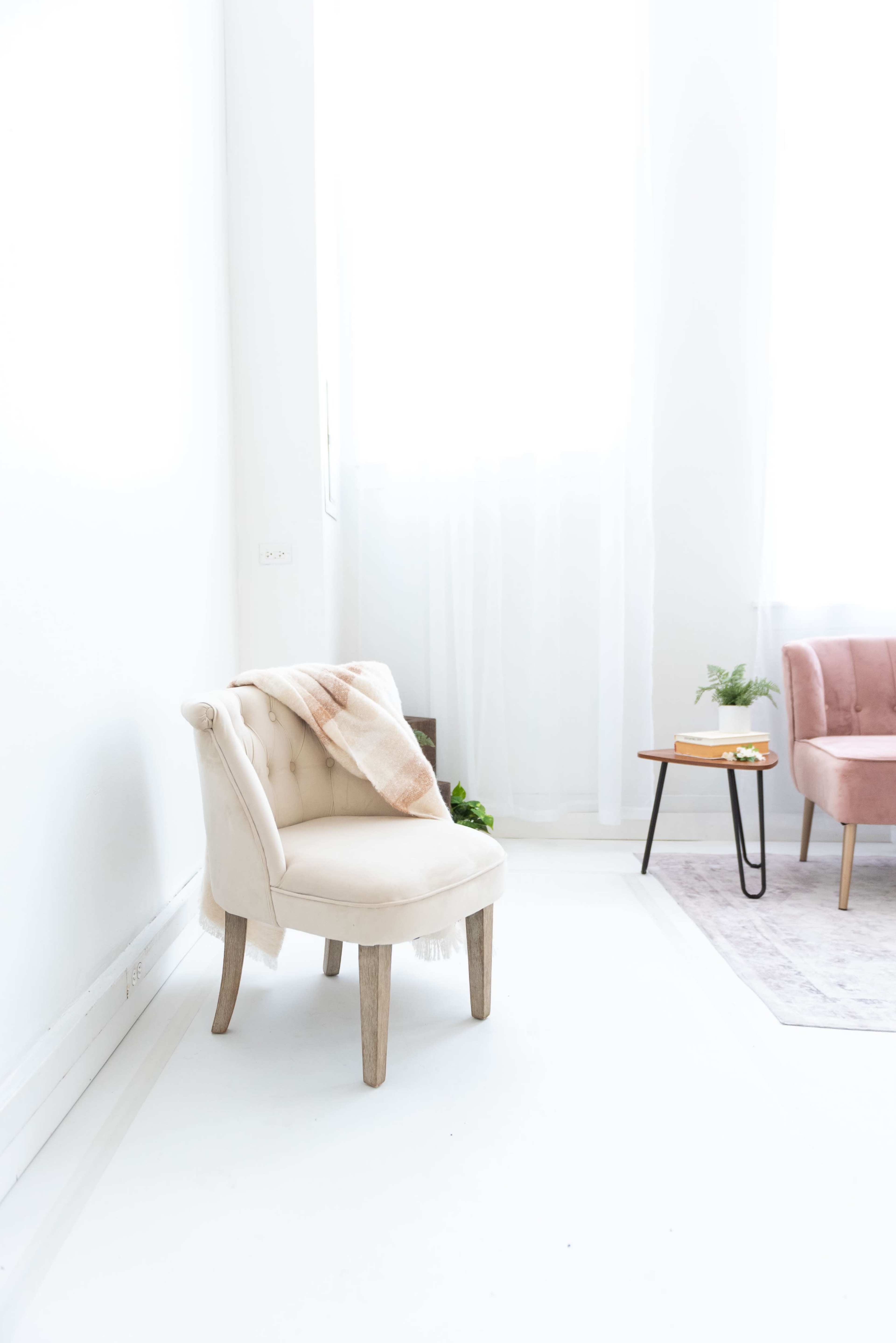 A light-colored chair with tufted upholstery and a blanket draped over it is placed next to a small table and another chair in a bright, minimalistic room.