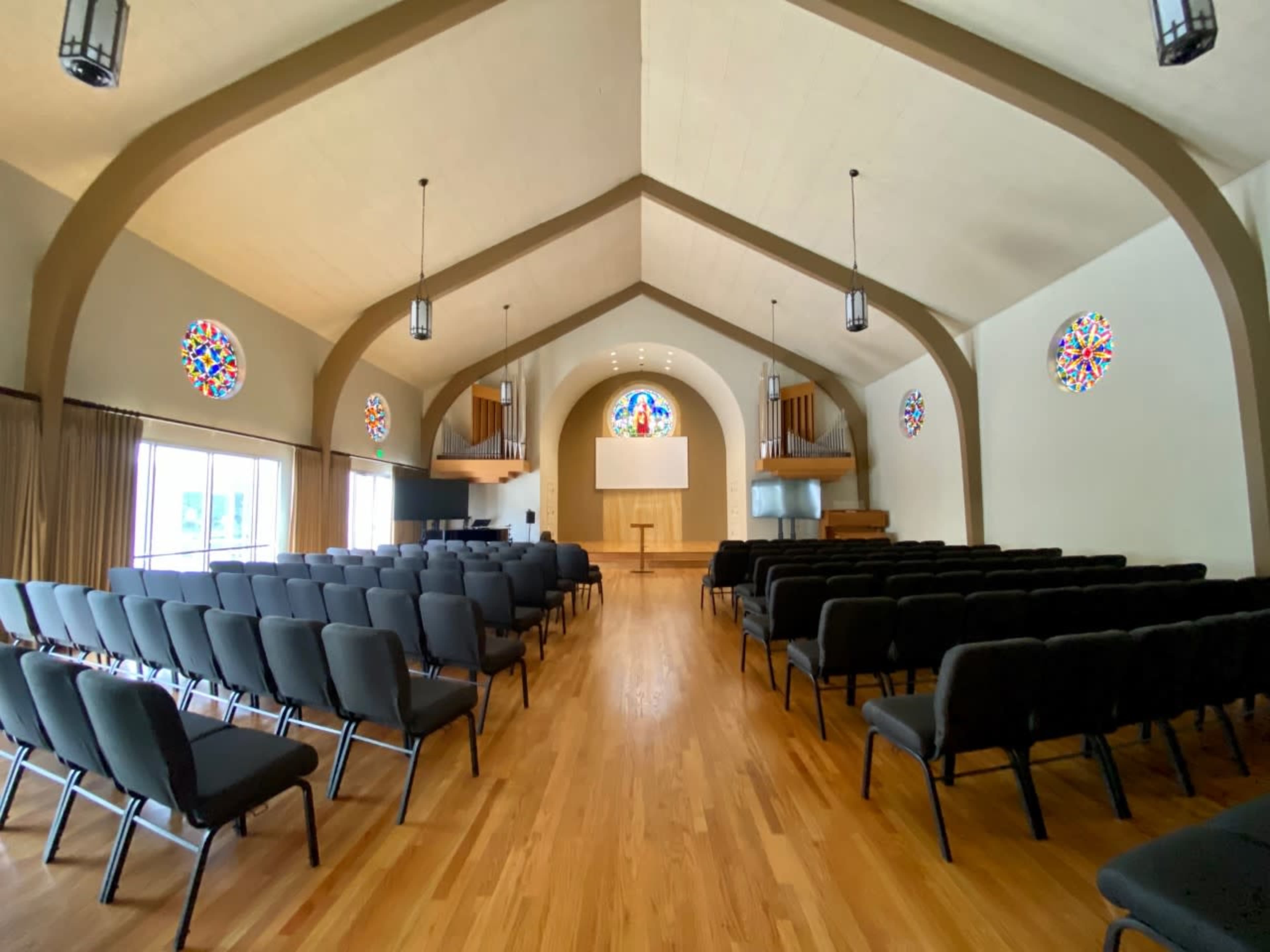 The interior of a church features rows of black chairs facing a wooden stage adorned with stained glass windows and wooden organ pipes.