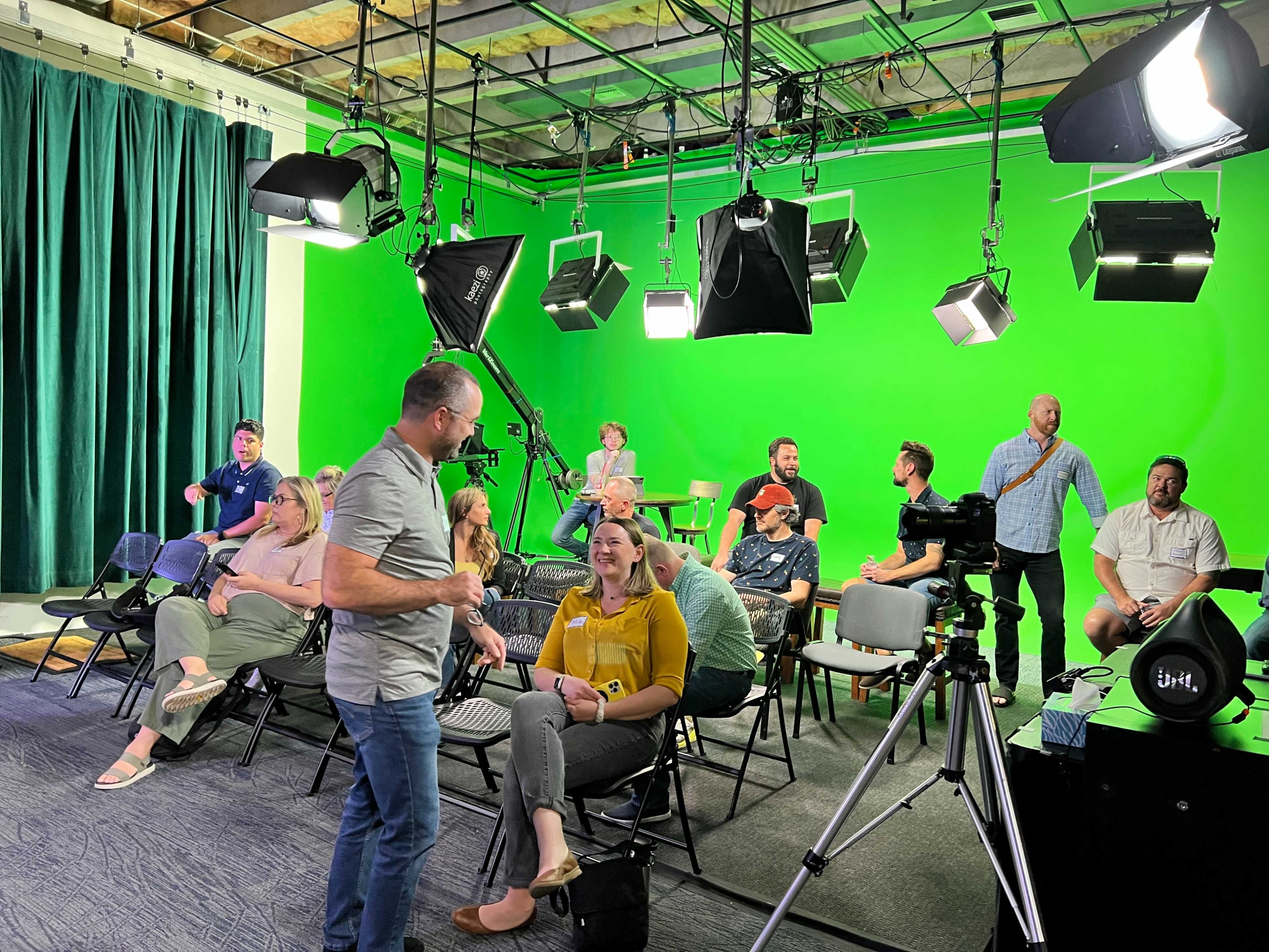 A group of people sits in chairs on a stage with a green screen backdrop, while a man stands and interacts with a woman.