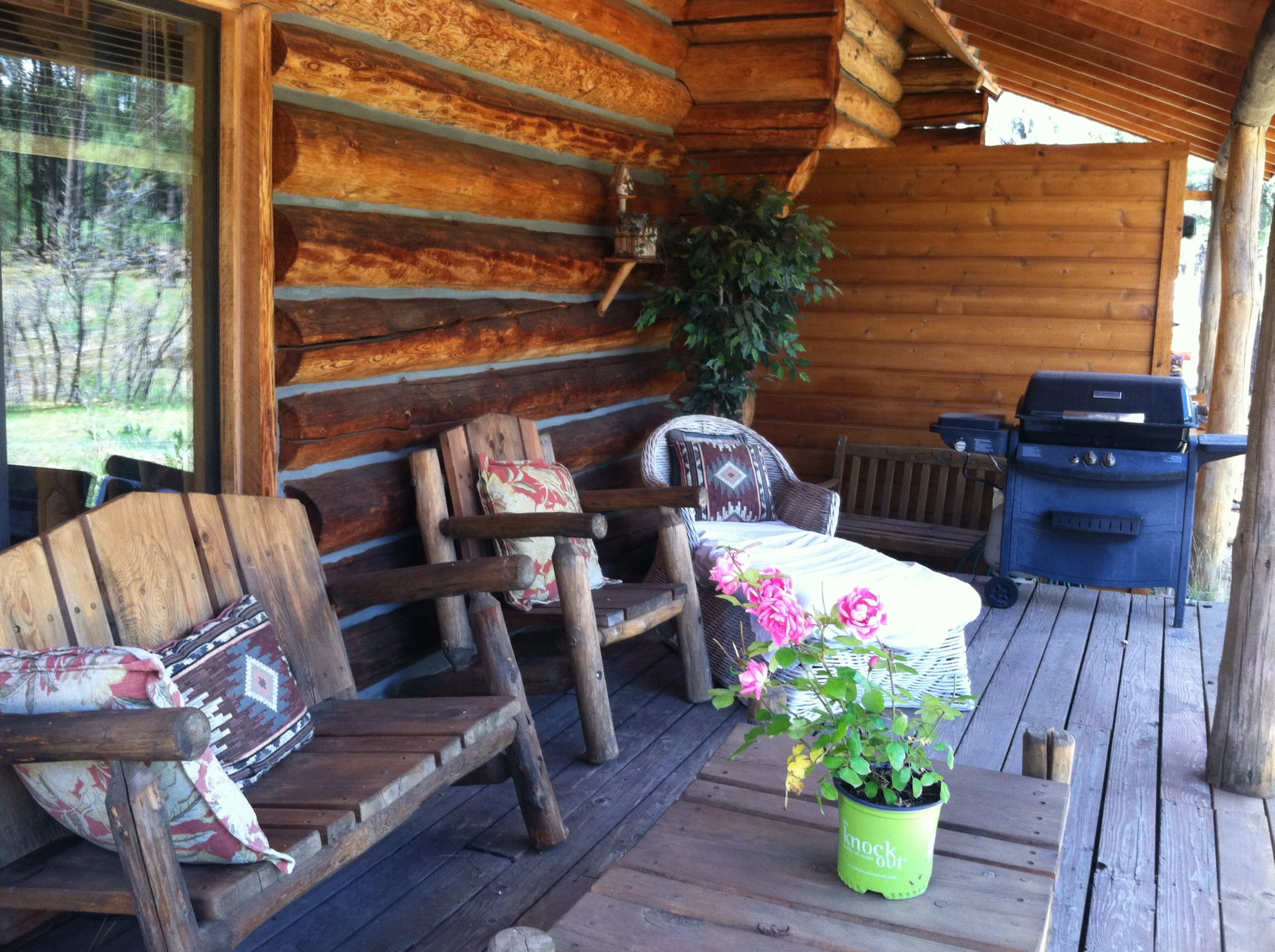 A wooden porch with two rustic chairs, a small table with a flower pot, and a grill in the background.