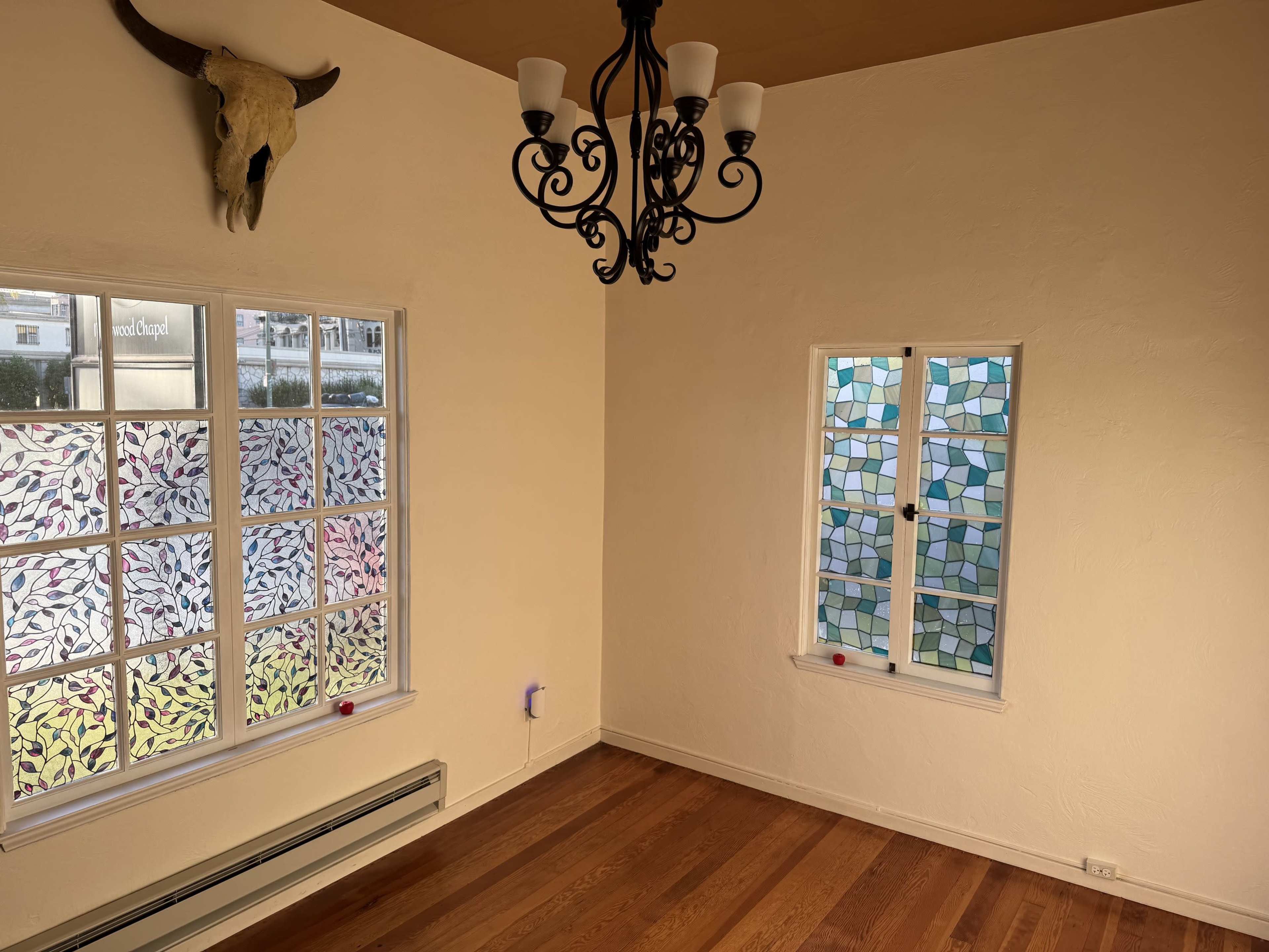 The image shows a room with wooden floors, light-colored walls, a stained glass window, and a chandelier hanging from the ceiling.