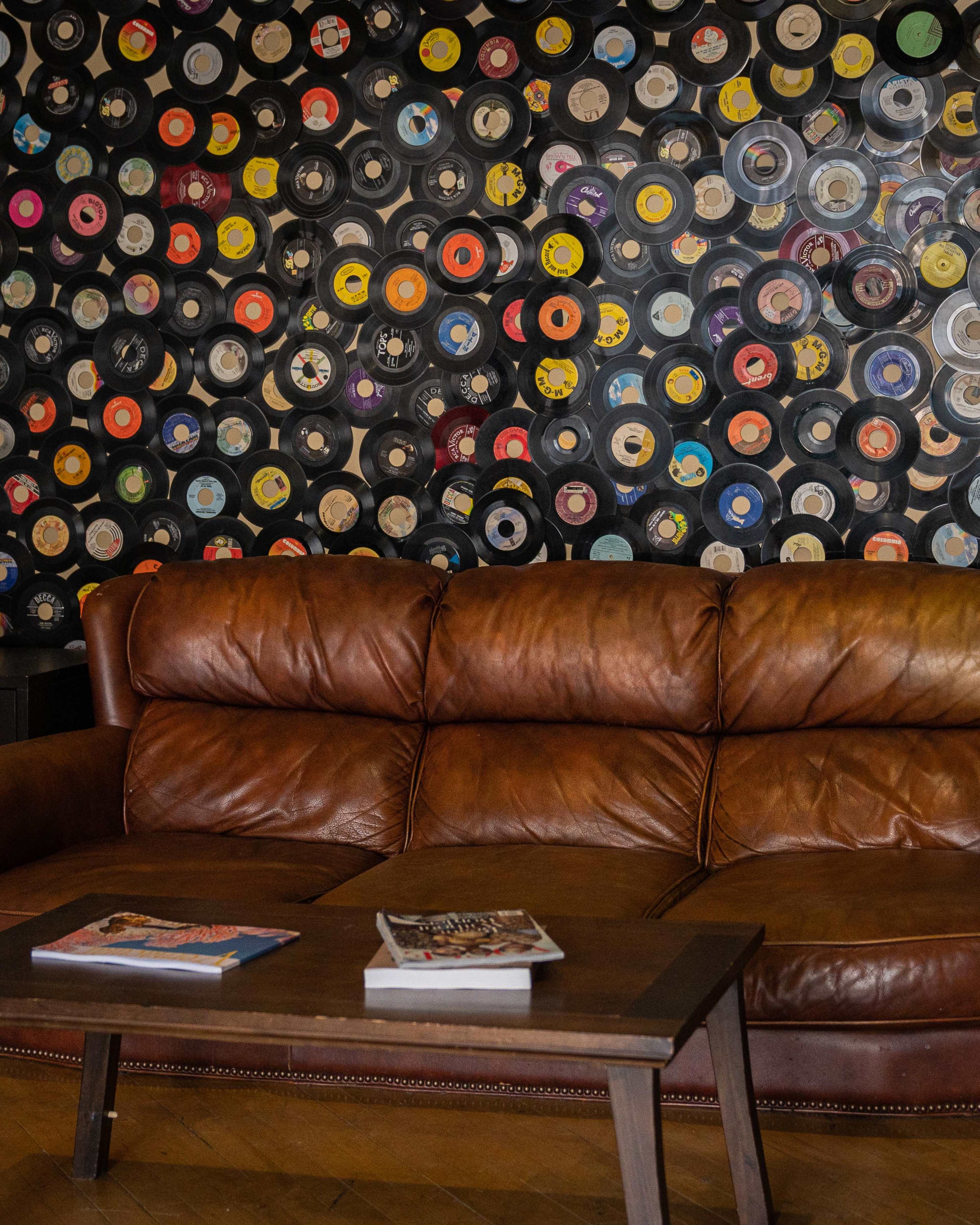 A brown leather couch is positioned in front of a colorful wall adorned with hanging vinyl records, while a wooden coffee table holds two magazines.
