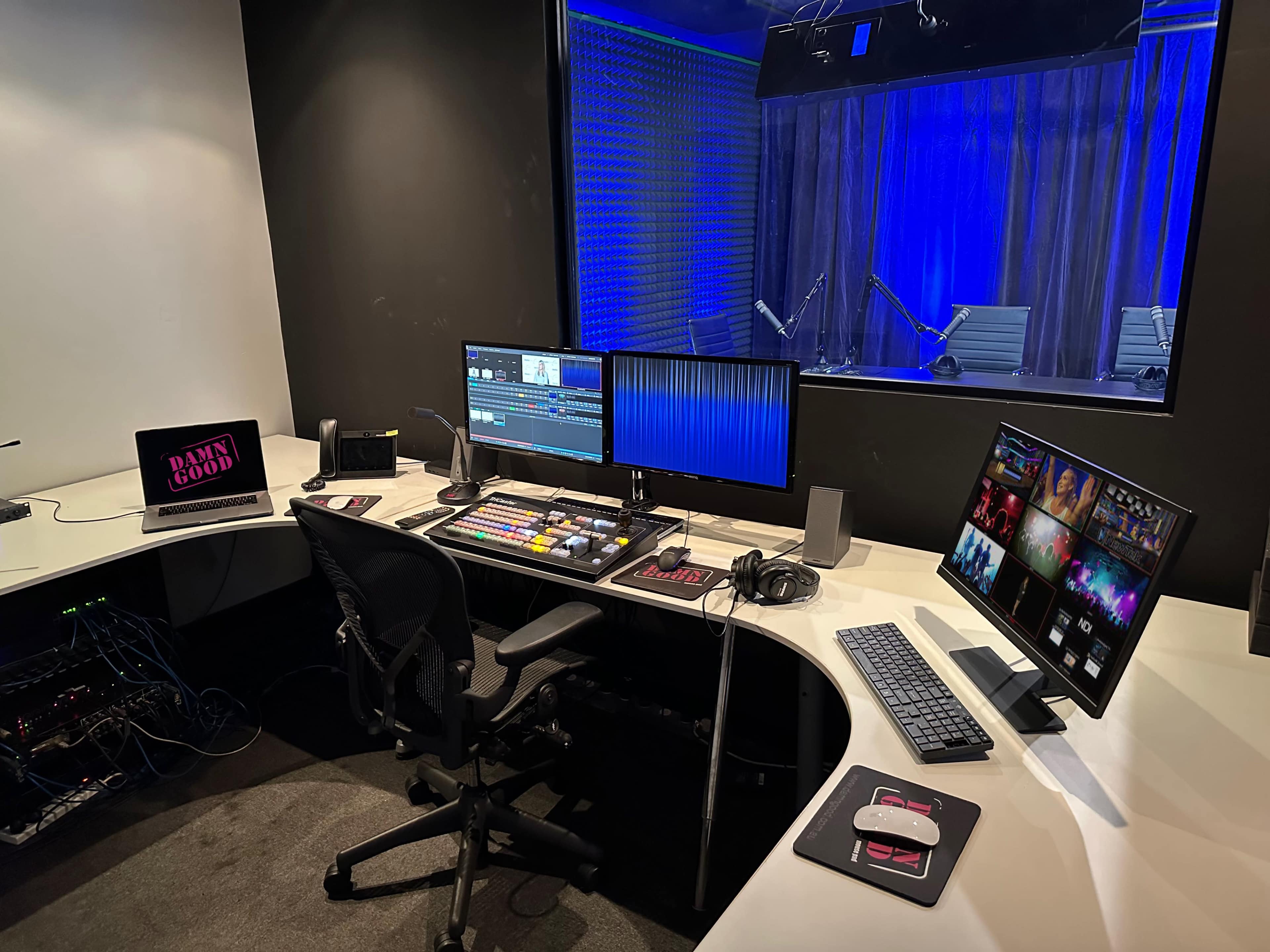 The image shows a modern control room with multiple computer screens, audio equipment, and a production console, set against a backdrop of blue curtains and lighting.