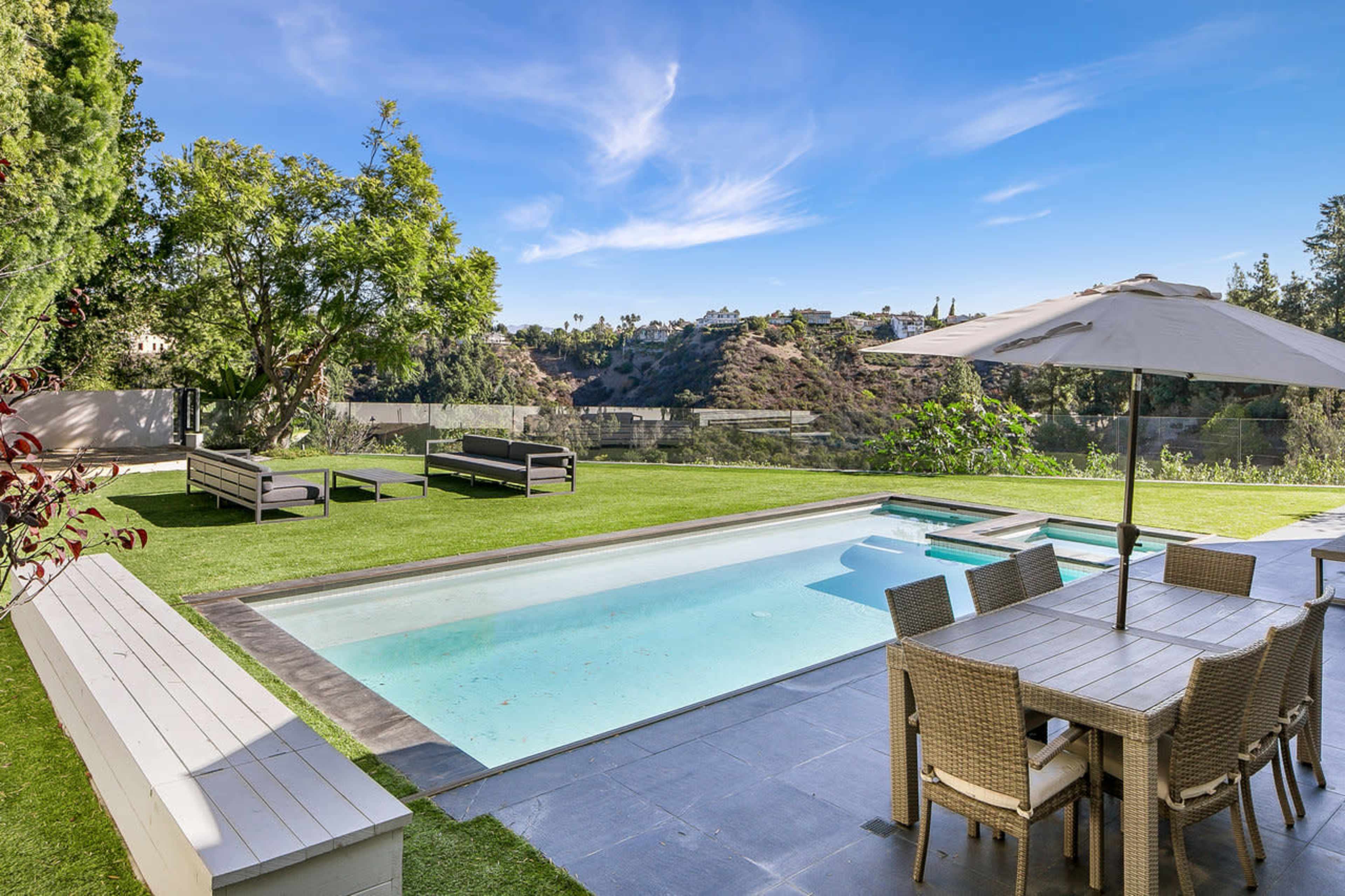 A pool area with lounge chairs and a dining table overlooks a hillside landscape.