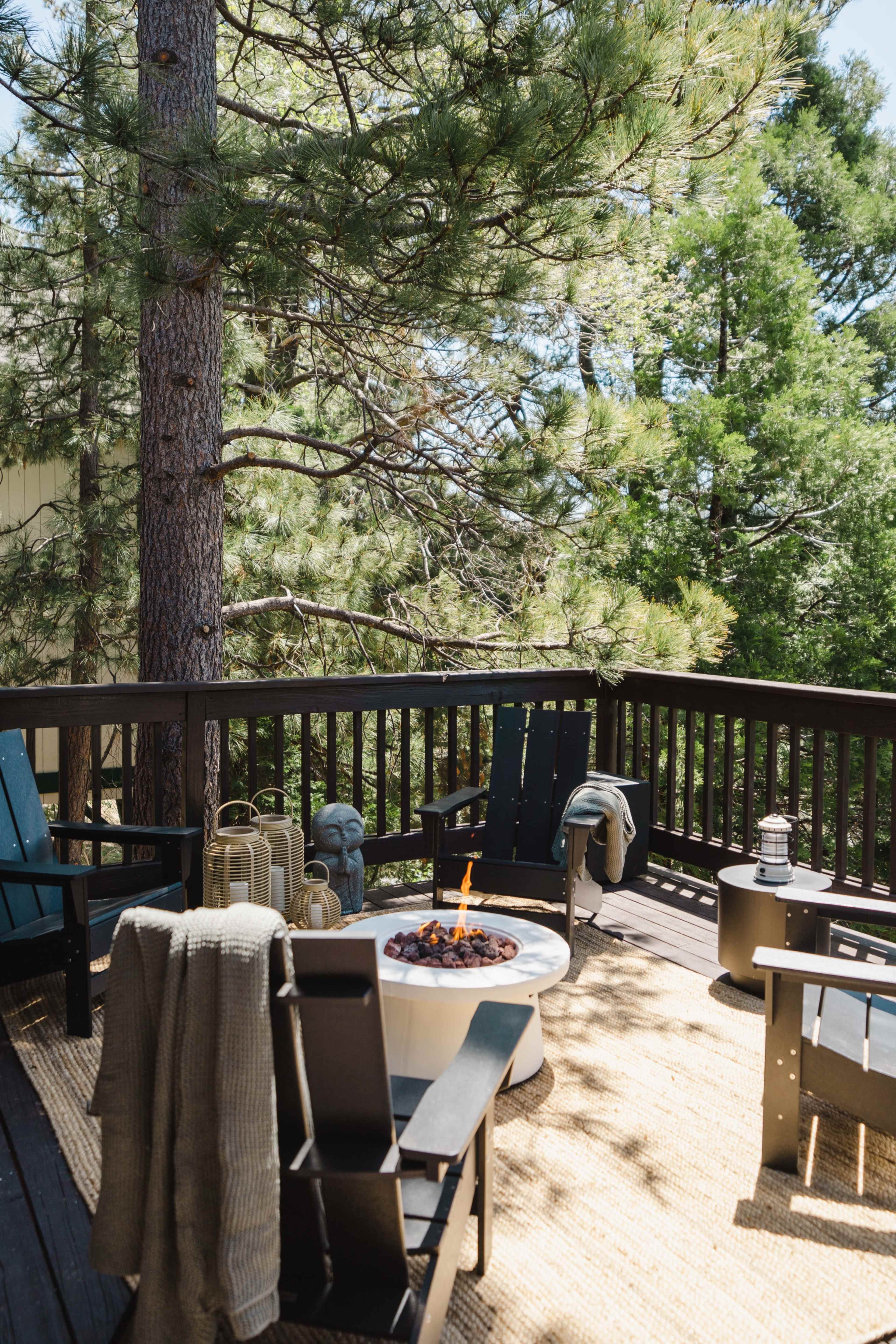 A wooden deck features black outdoor chairs arranged around a fire pit, surrounded by tall trees.