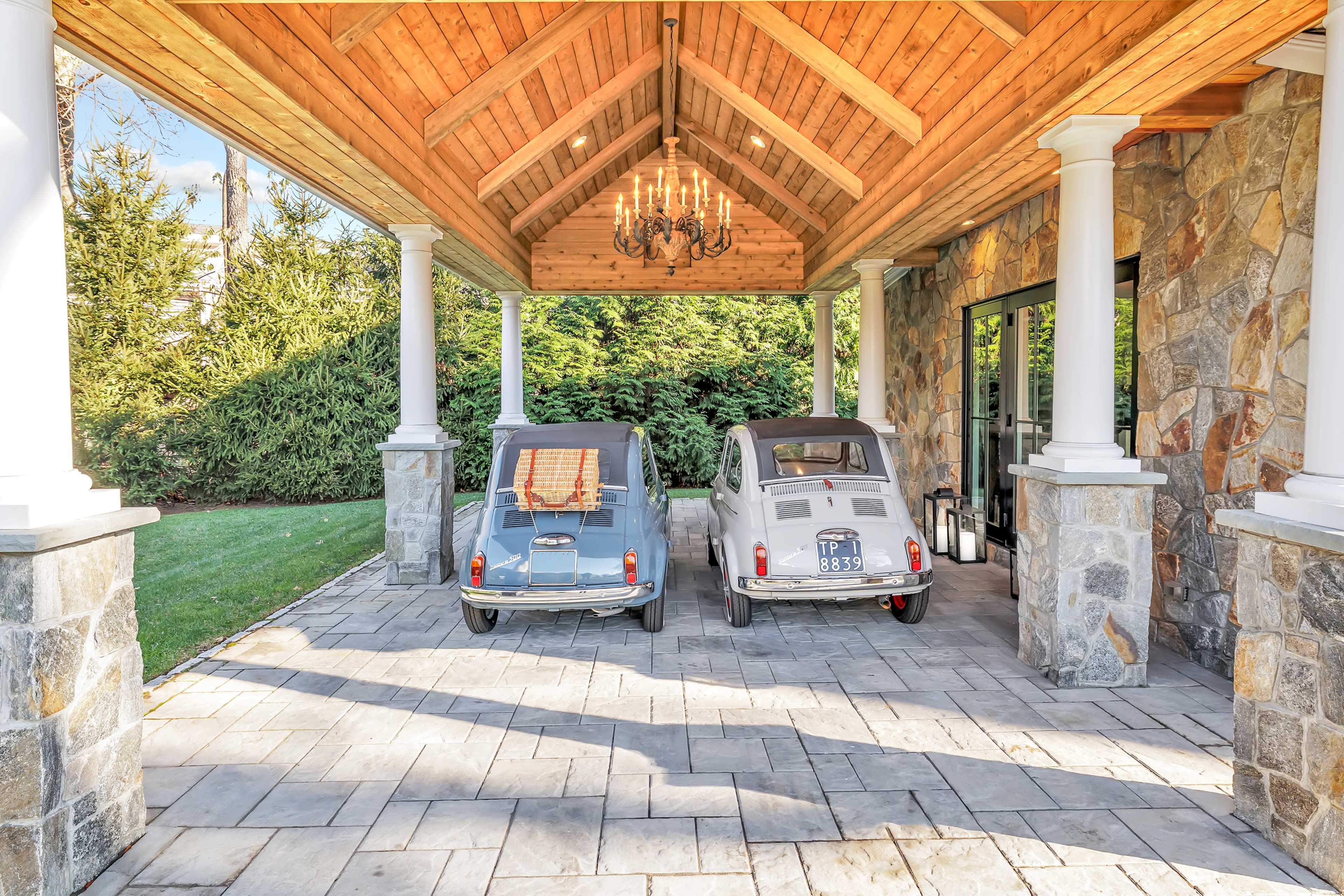 Two vintage cars are parked under a wooden-beamed portico adorned with a chandelier, next to a stone wall, on a paved area surrounded by greenery.