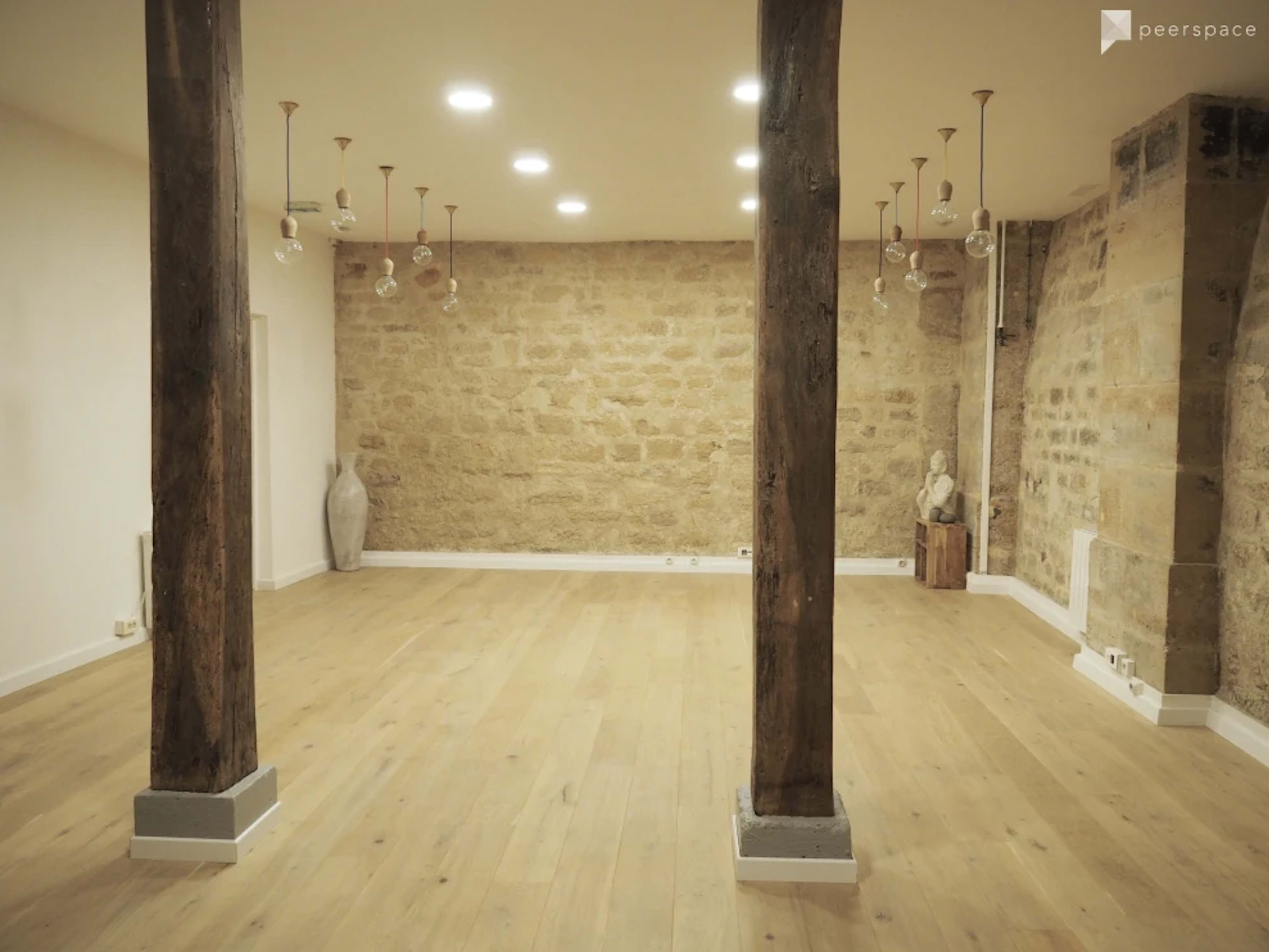 A spacious room with wooden flooring, exposed stone walls, and wooden support beams, illuminated by pendant lights.