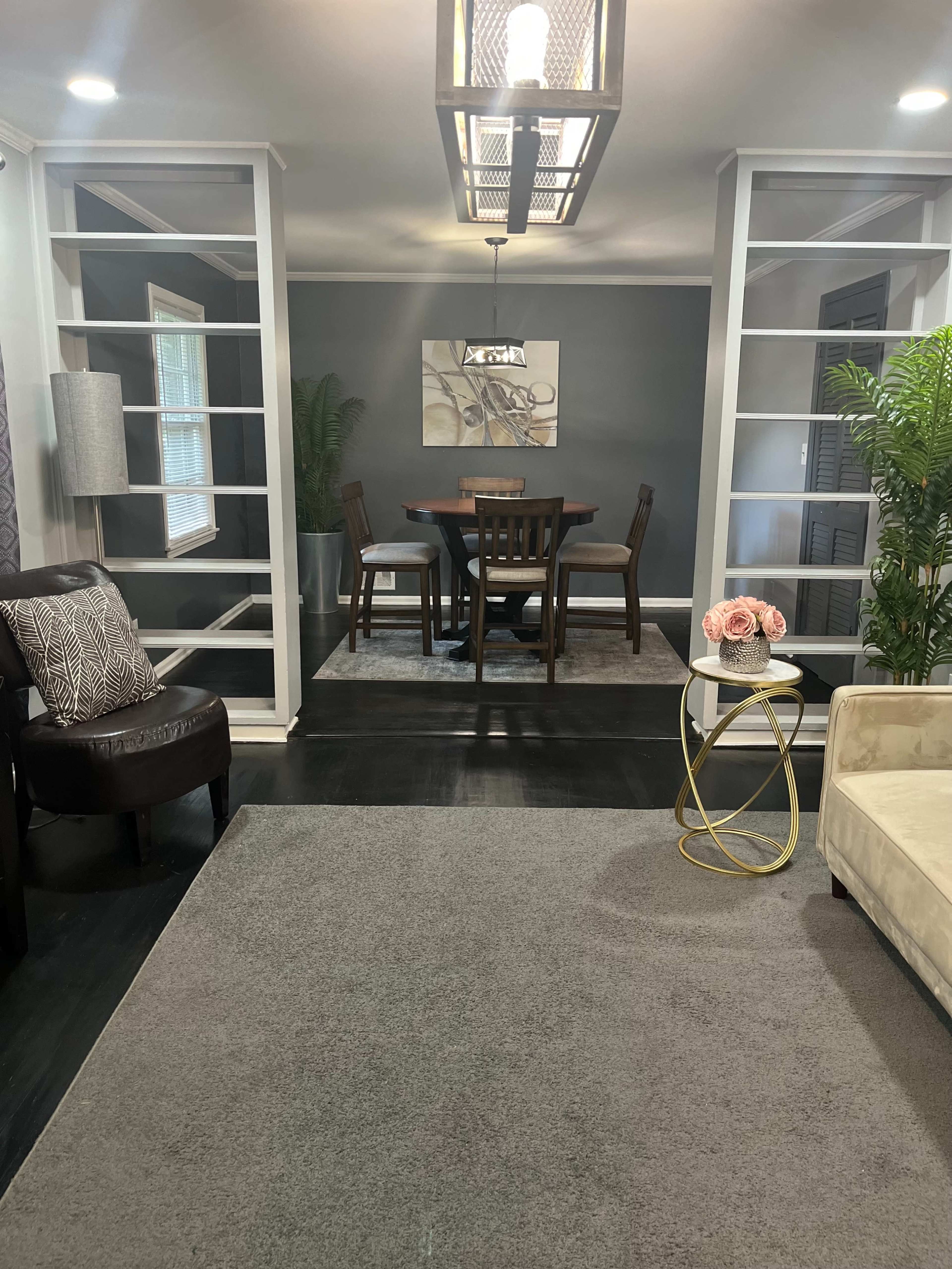 The image shows a well-decorated dining area with a round table and four chairs, framed by glass partitions and complemented by a cozy seating area with a chair and side table.