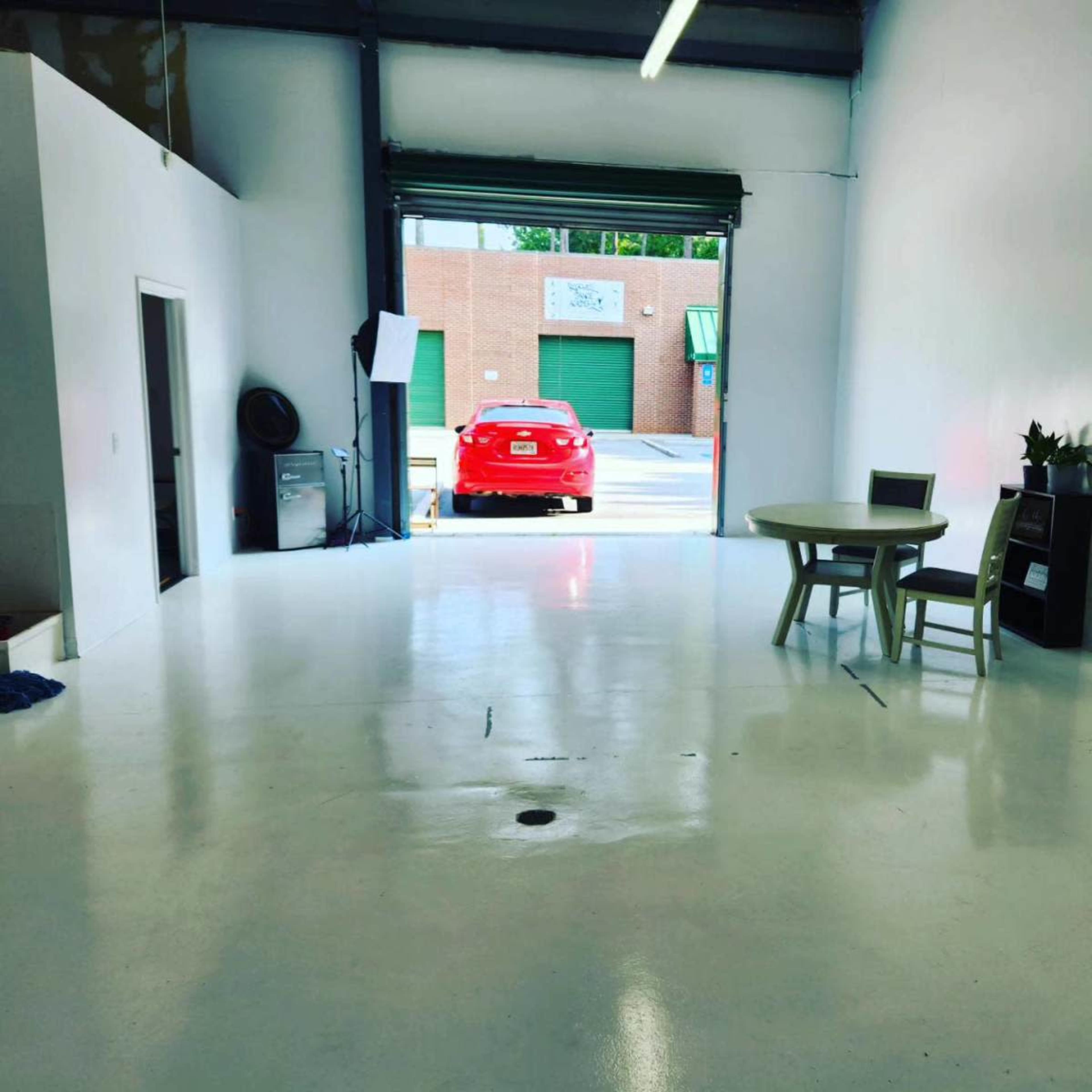 The image depicts a bright, spacious room with a white floor, a table and chairs, and an open garage door revealing a red car parked outside.