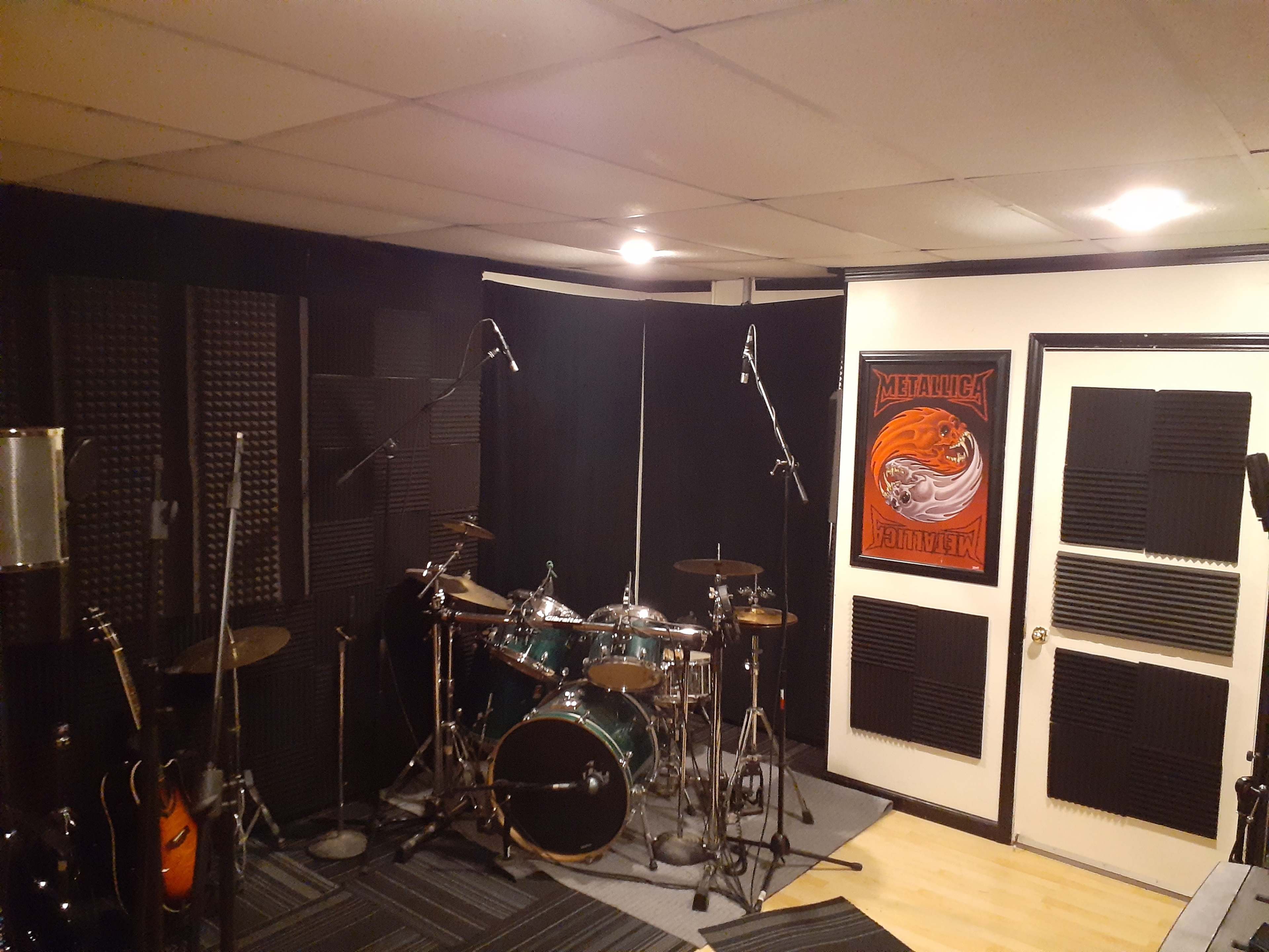 A music practice room featuring a drum set, electric guitars, microphones on stands, and soundproofing panels on the walls.