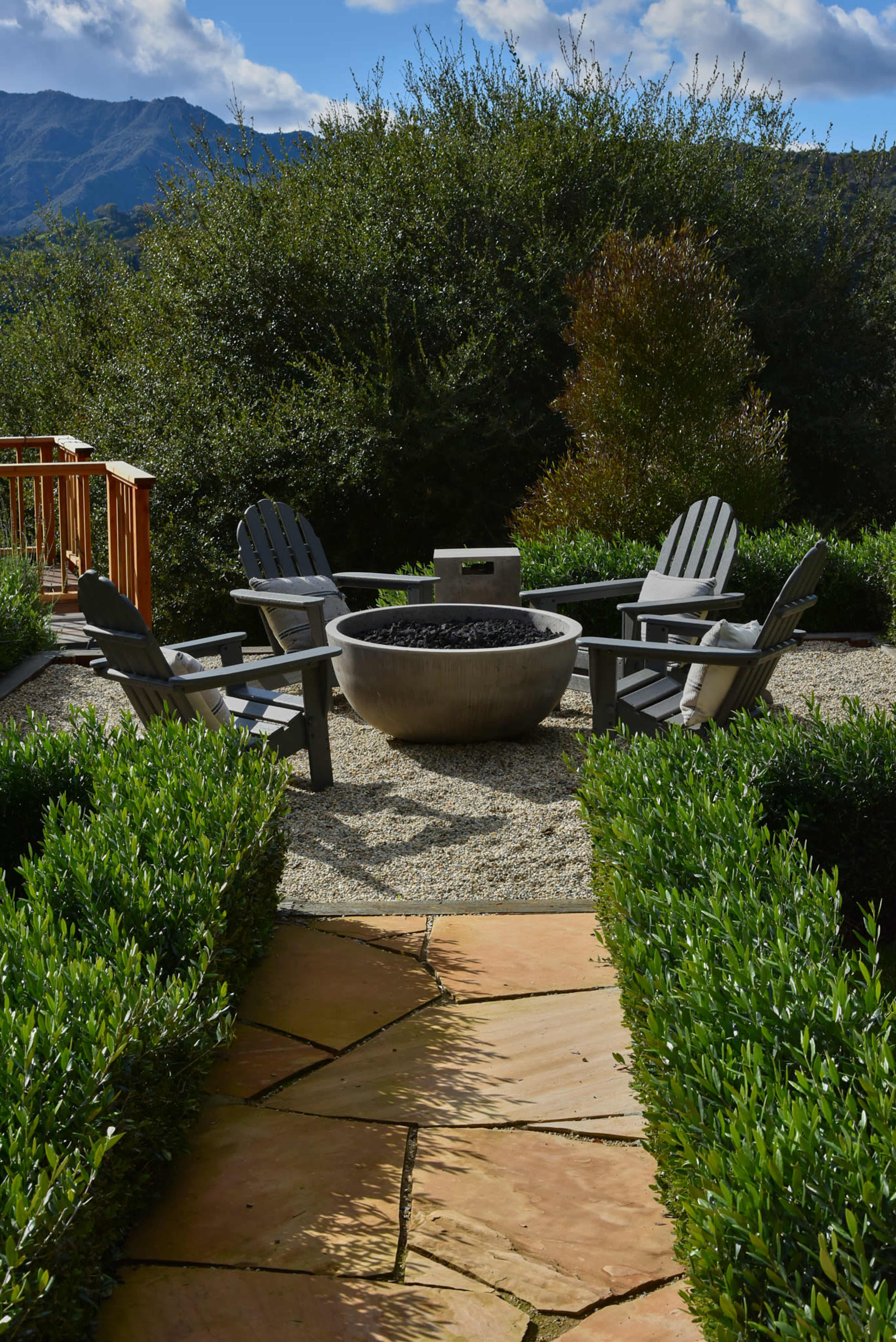A circular stone fire pit surrounded by four wooden chairs is situated in a landscaped garden with shrubs and a backdrop of mountains.