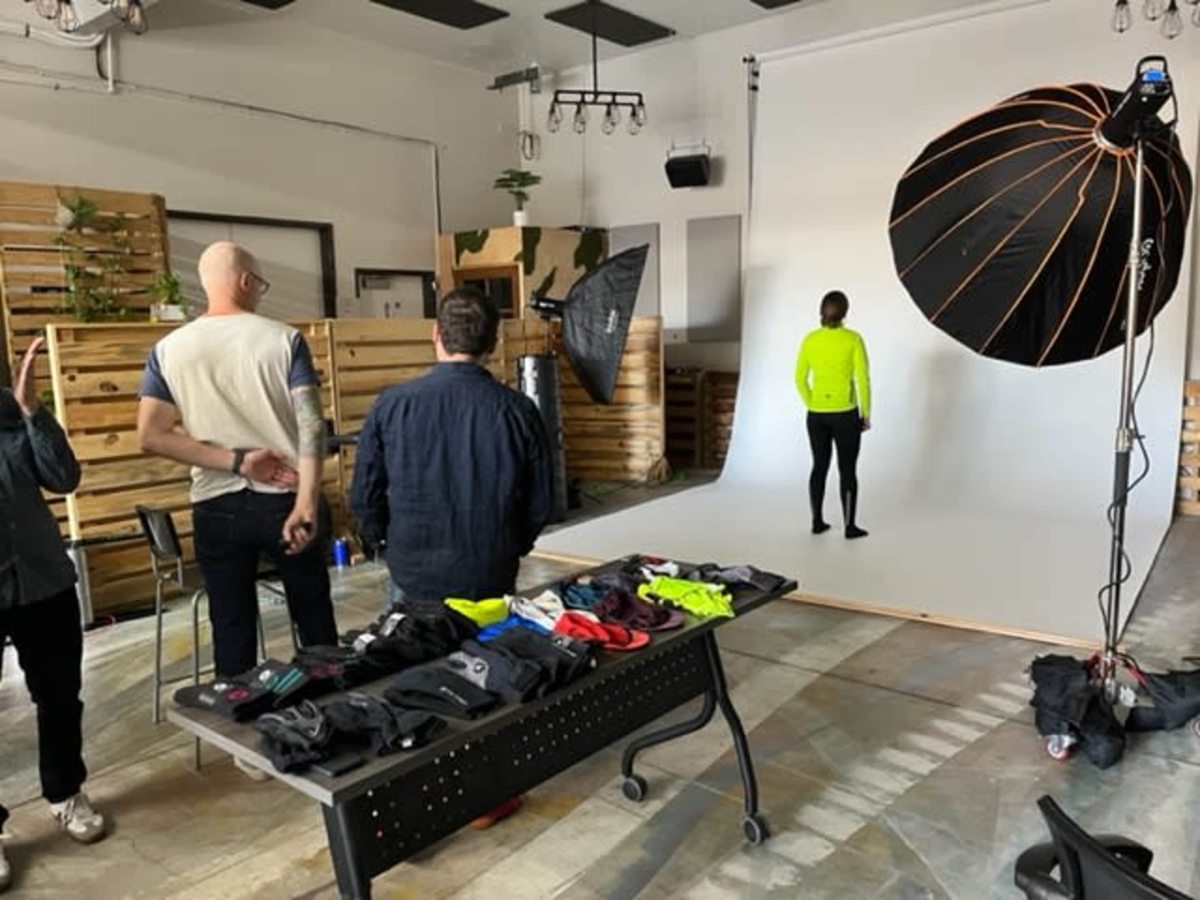 A photography studio setup features a model in a bright outfit posing against a neutral background, while crew members arrange equipment and clothing on a table.