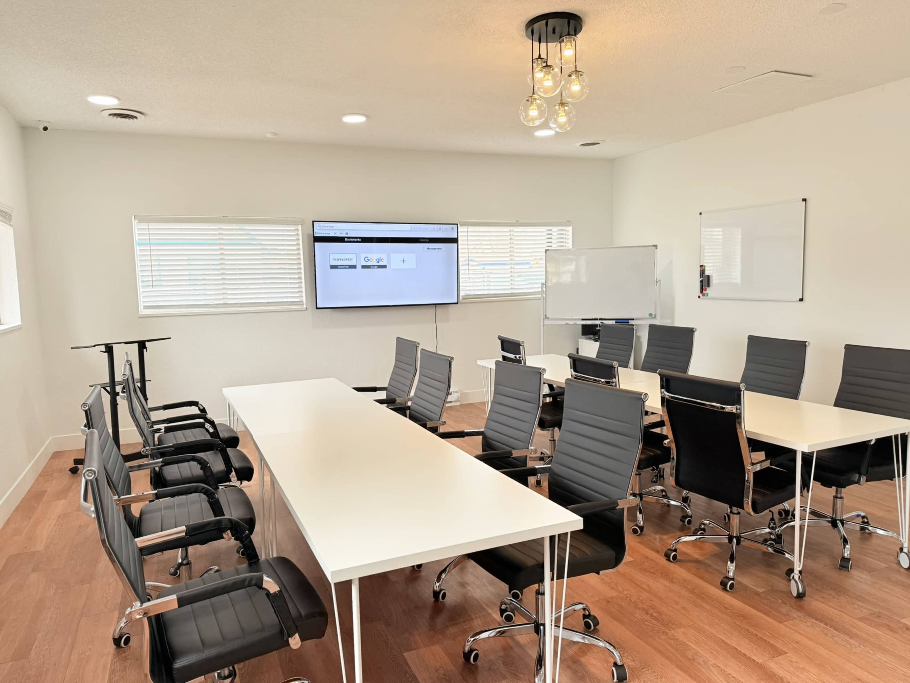 The image shows a modern conference room with a long white table, black office chairs, a large screen on the wall, and whiteboards.