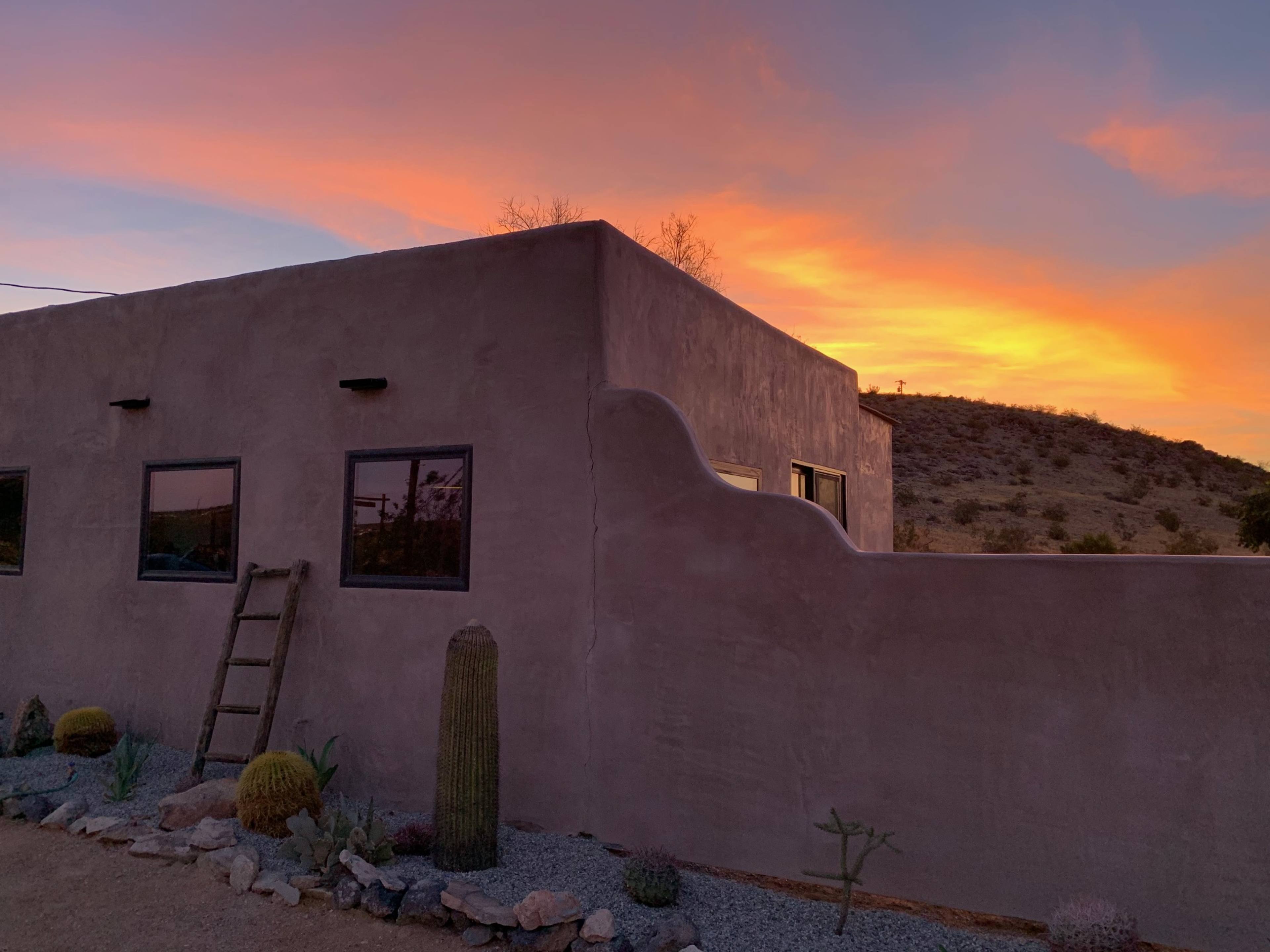 A stucco building with large windows is set against a vibrant sunset sky, with a cactus and a wooden ladder leaning against the wall.