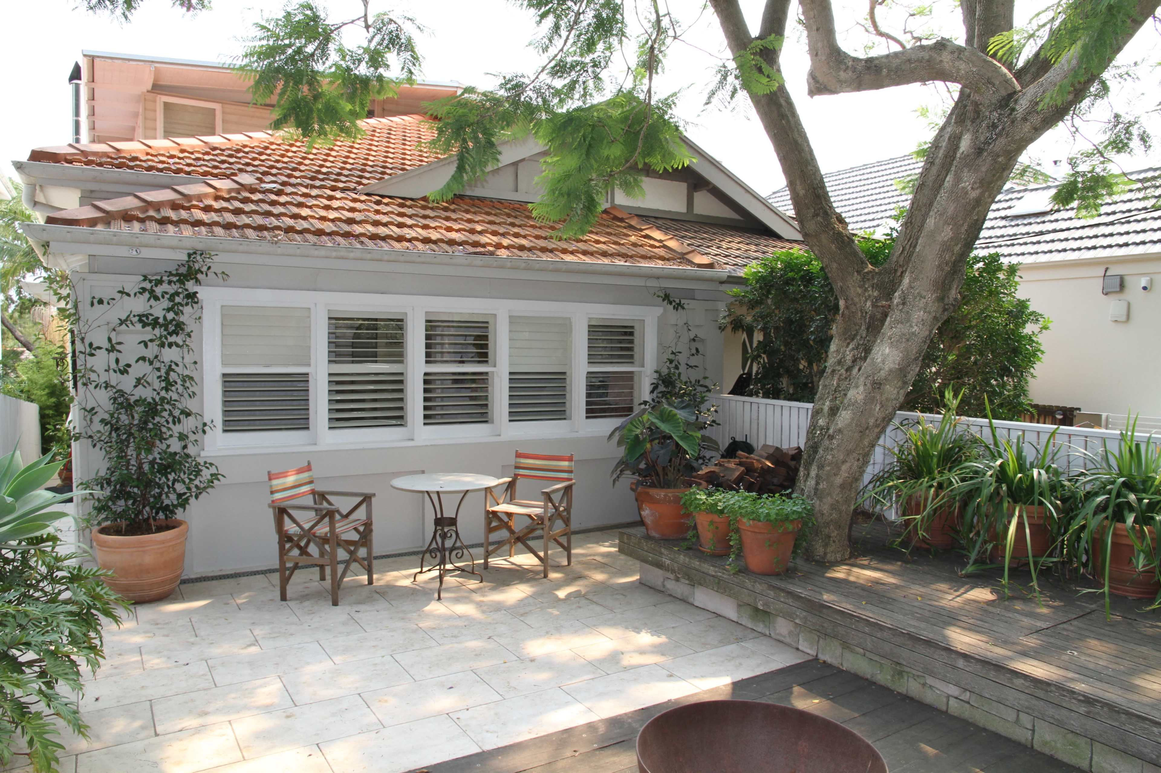 A patio area features two chairs and a small table beside a house with a tiled roof, surrounded by potted plants and a large tree.