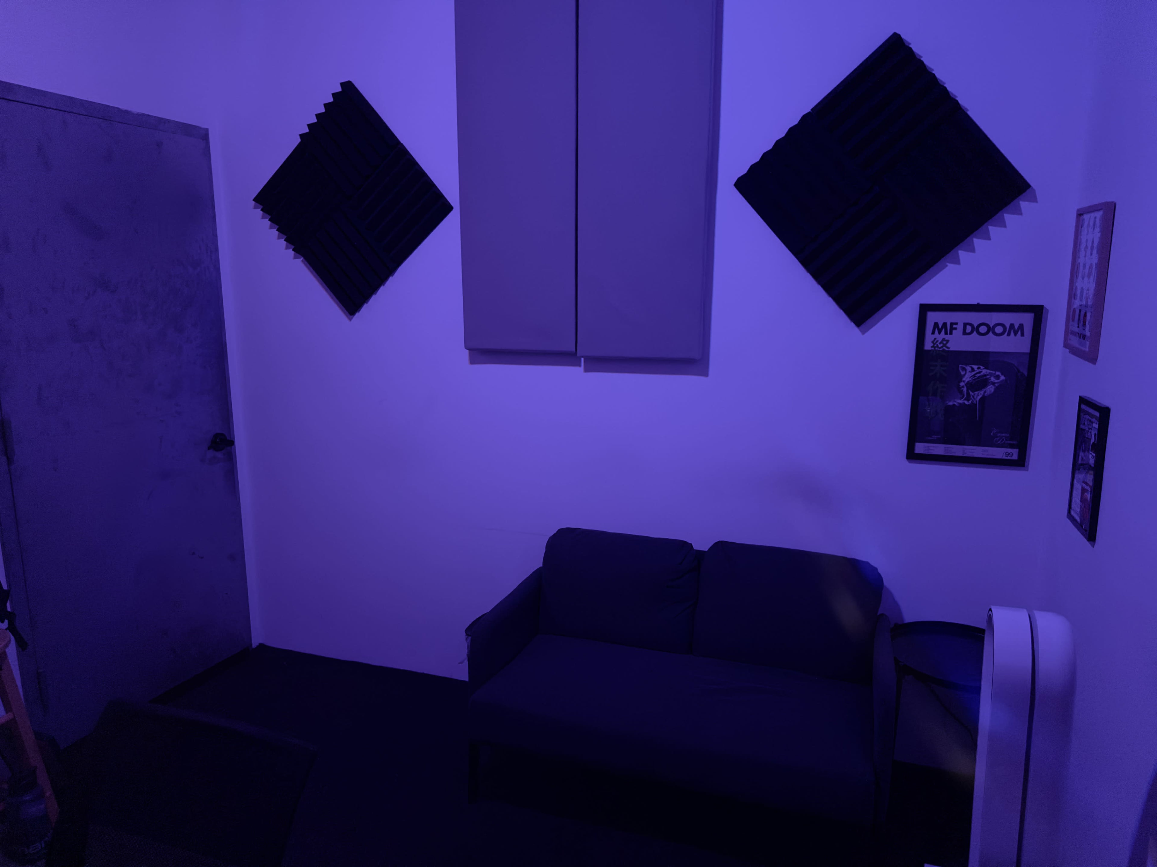 The image shows a small room with a dark couch, soundproofing panels on the walls, and a poster framed on the wall.