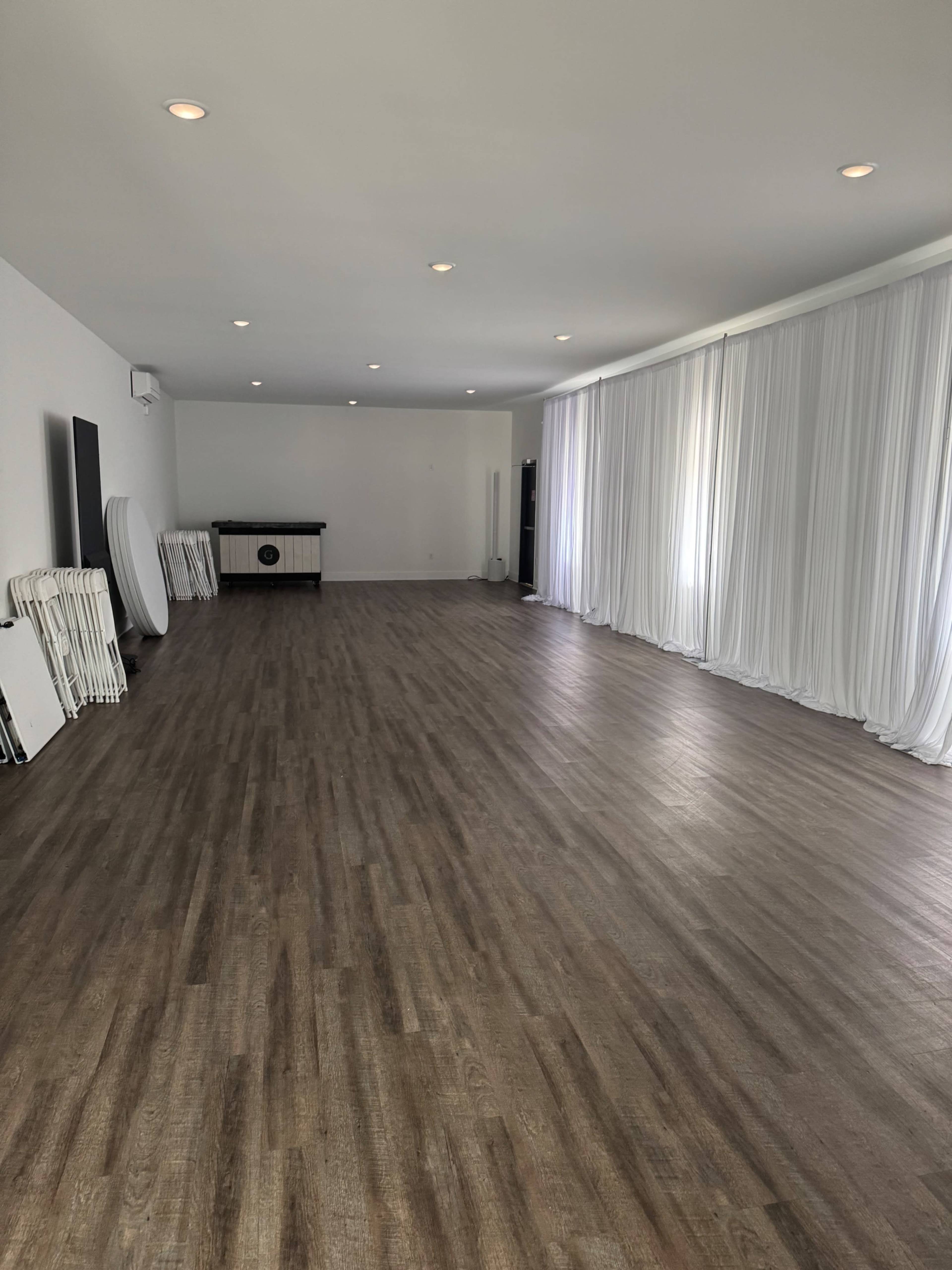 The image shows a spacious interior room with wooden floors, white draped curtains, and a few tables stacked against one wall.