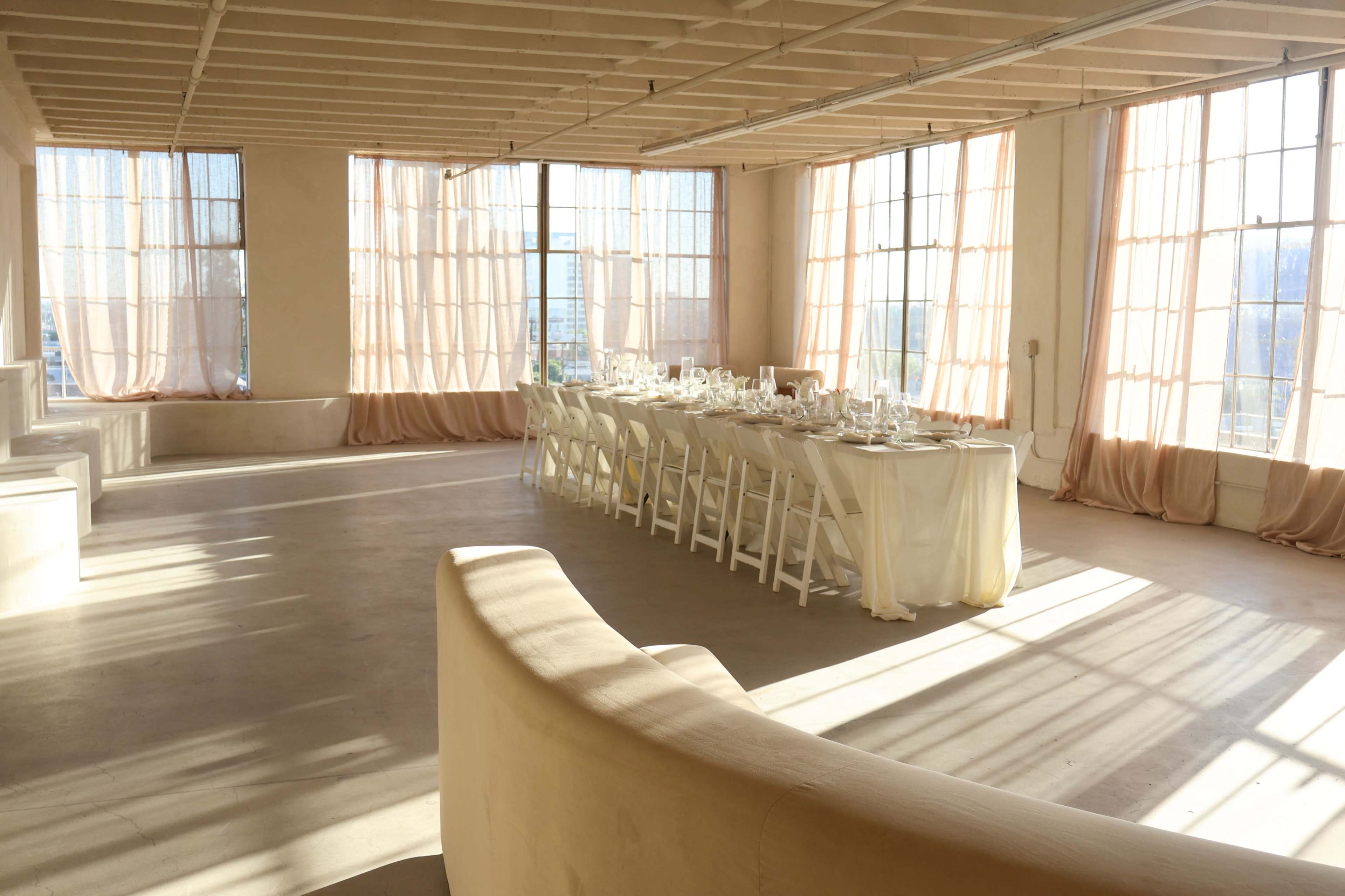 A long dining table is set with glassware and white table linens in a spacious, well-lit room with large windows and soft drapes.