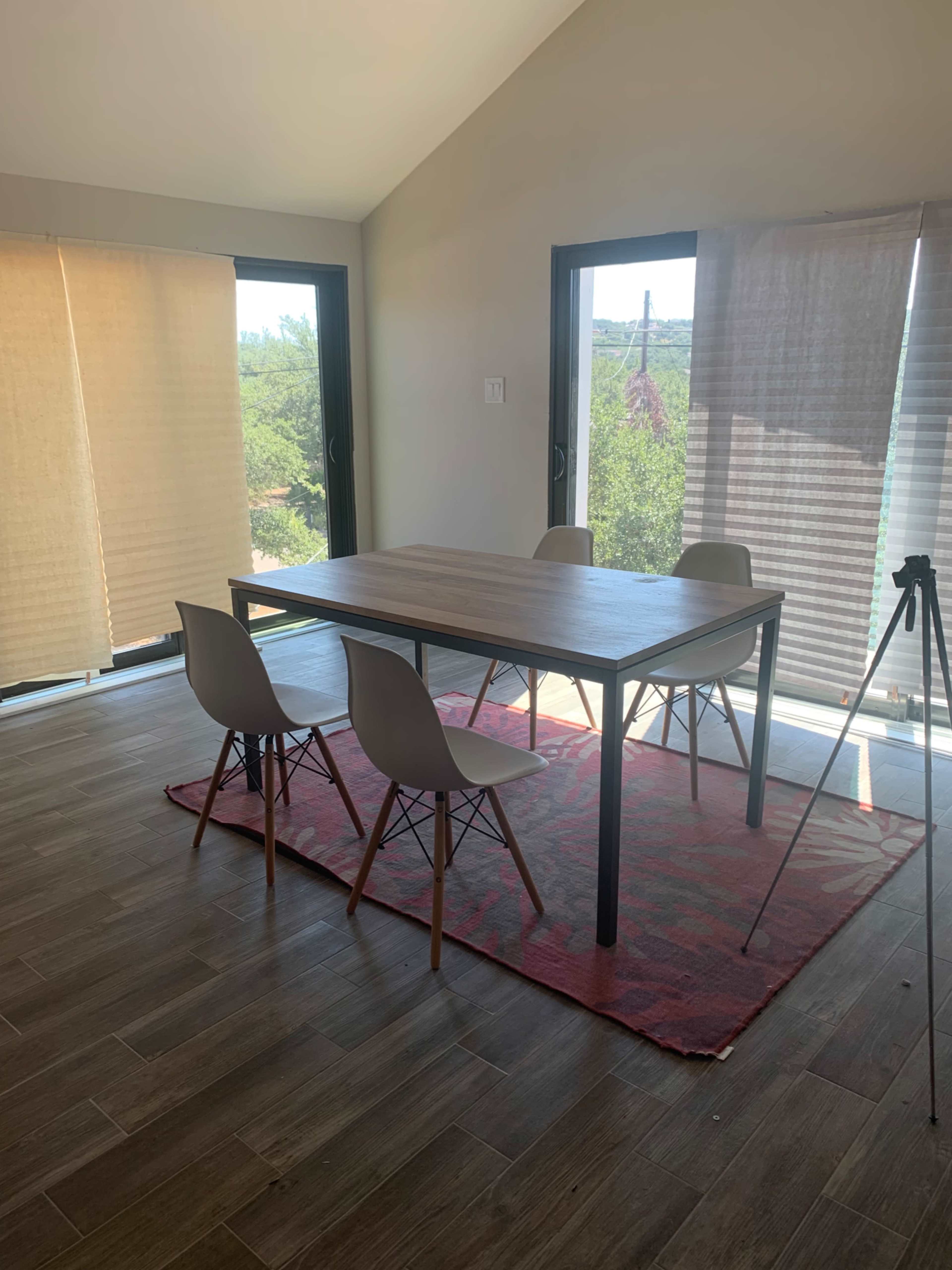 A wooden table with four chairs is positioned on a red rug in a bright room featuring large windows with blinds.