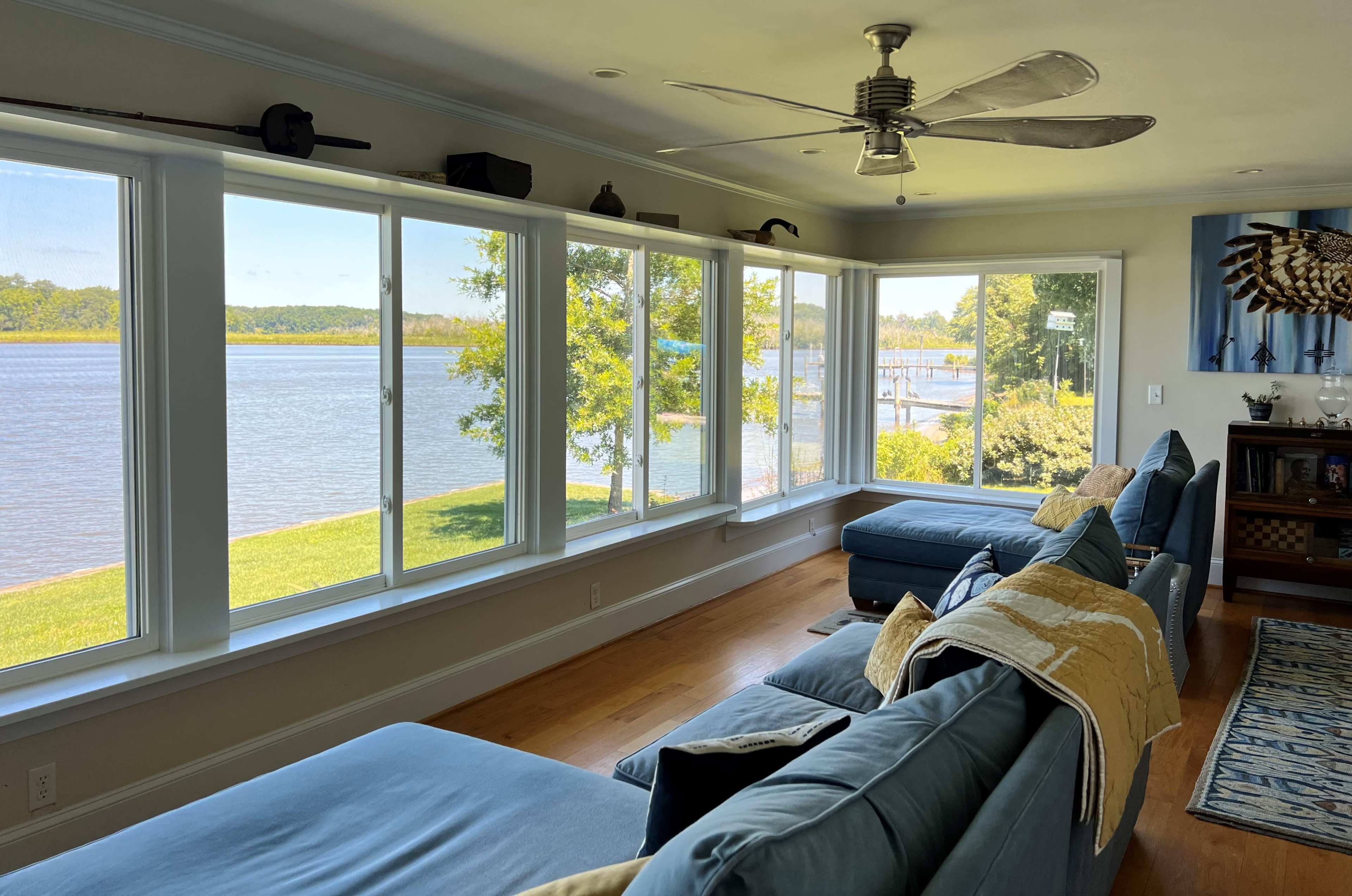 A spacious living area features large windows overlooking a river and green landscape, with blue sofas and a ceiling fan.