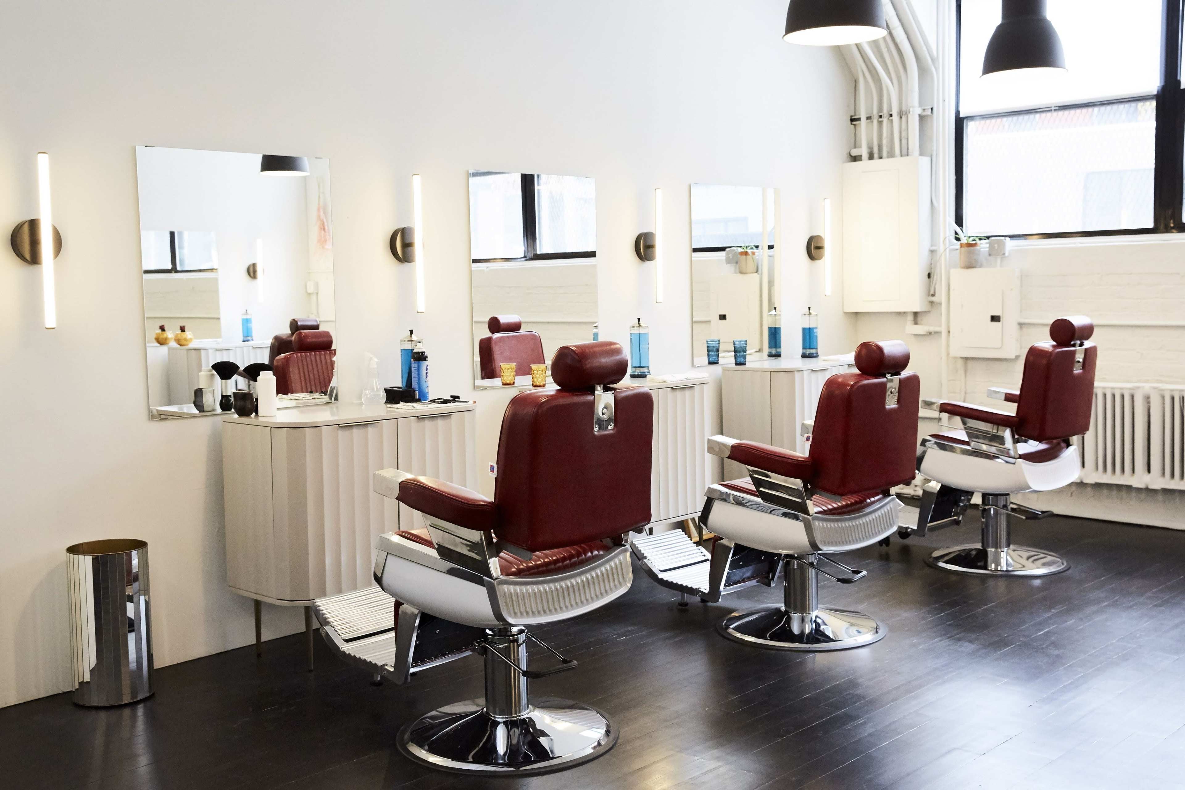A modern hair salon with three red barber chairs facing mirrors and several grooming products displayed on a white counter.