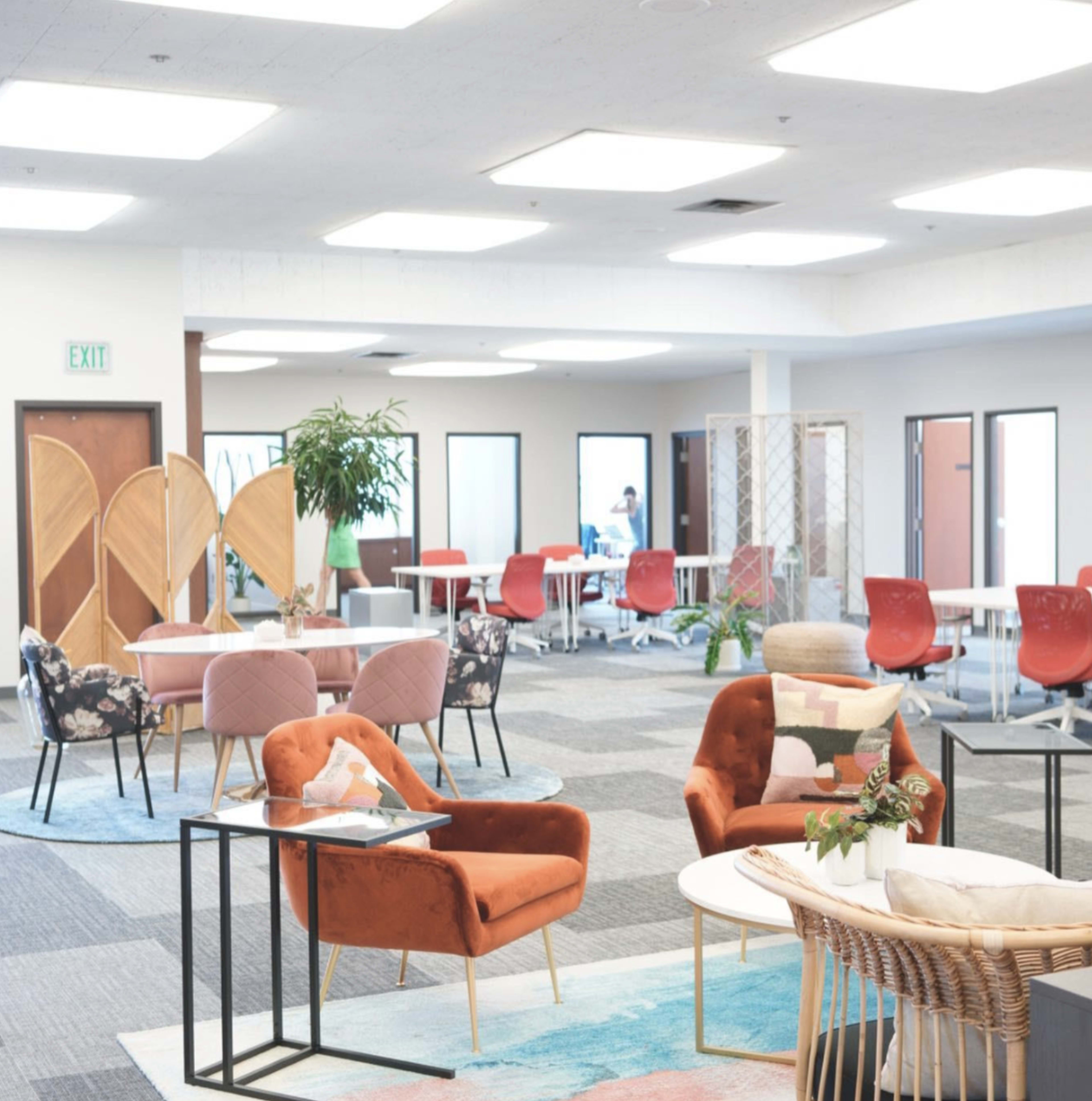 The image shows a modern, open workspace with various seating areas, tables, and a blend of colorful furniture arranged across the carpeted floors.