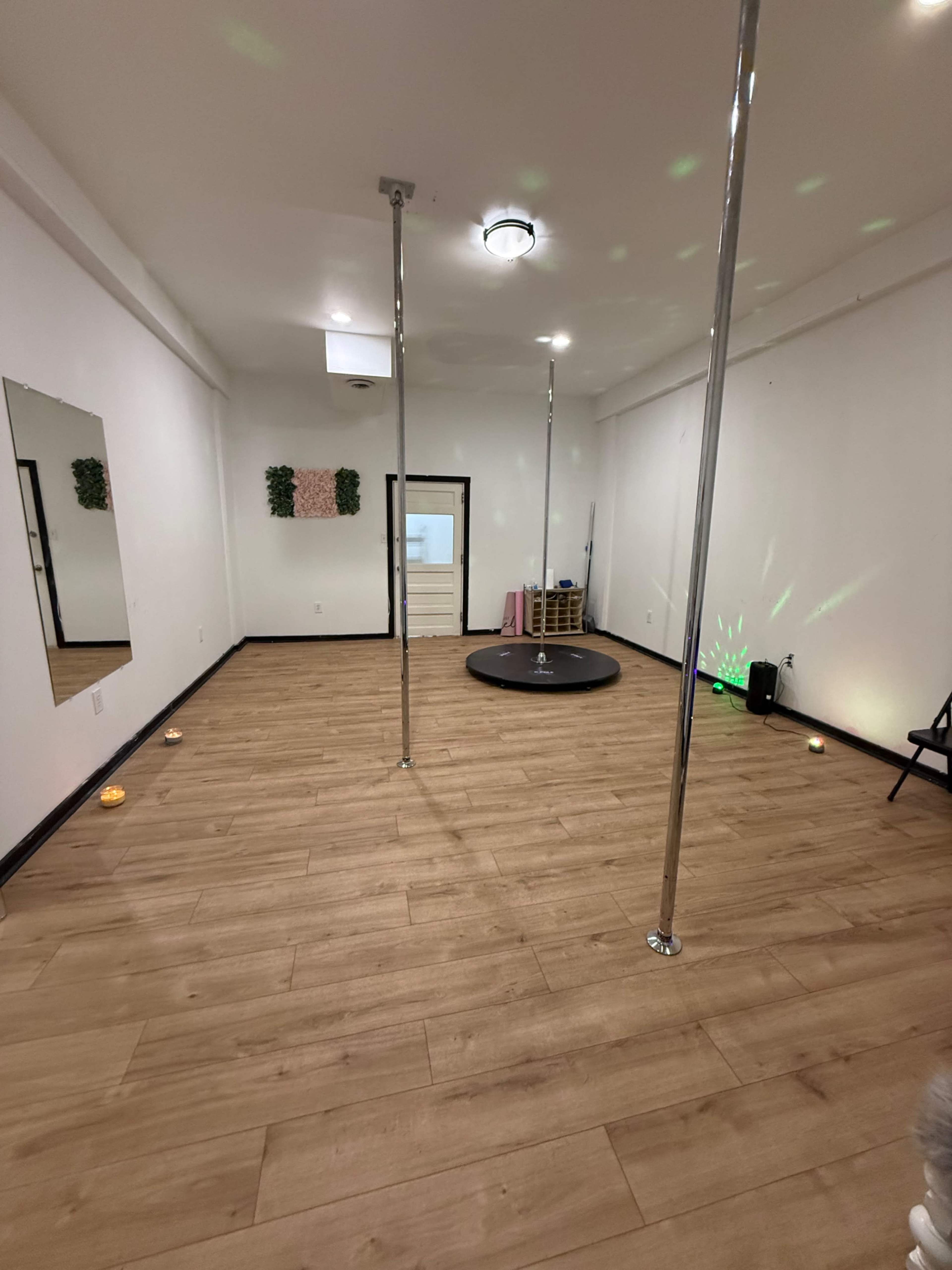 The image shows a spacious dance studio with wooden flooring, two vertical poles, a circular stage, and soft lighting elements.