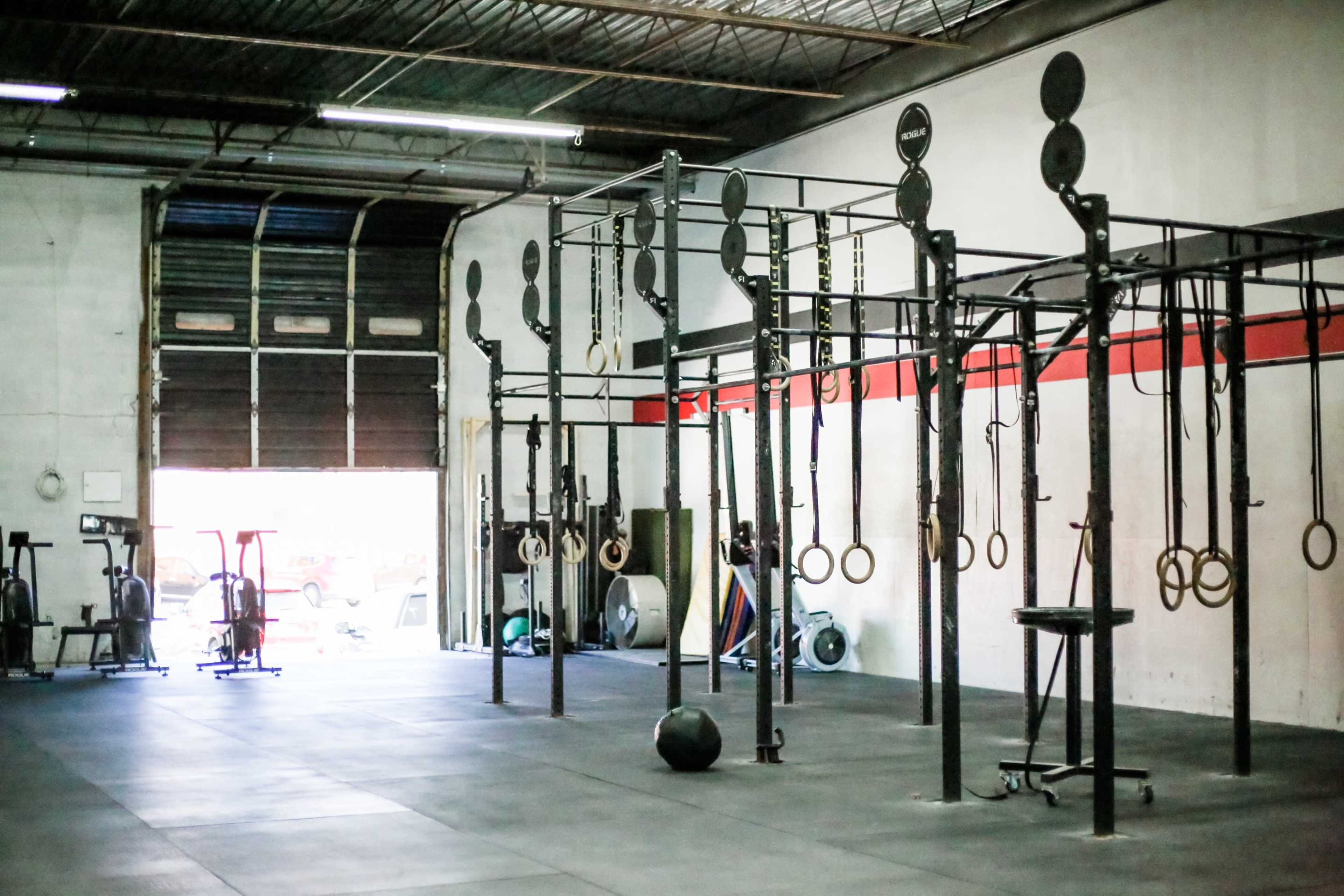A gym interior with various workout equipment, including cable machines, rings, and a medicine ball, against a concrete wall.