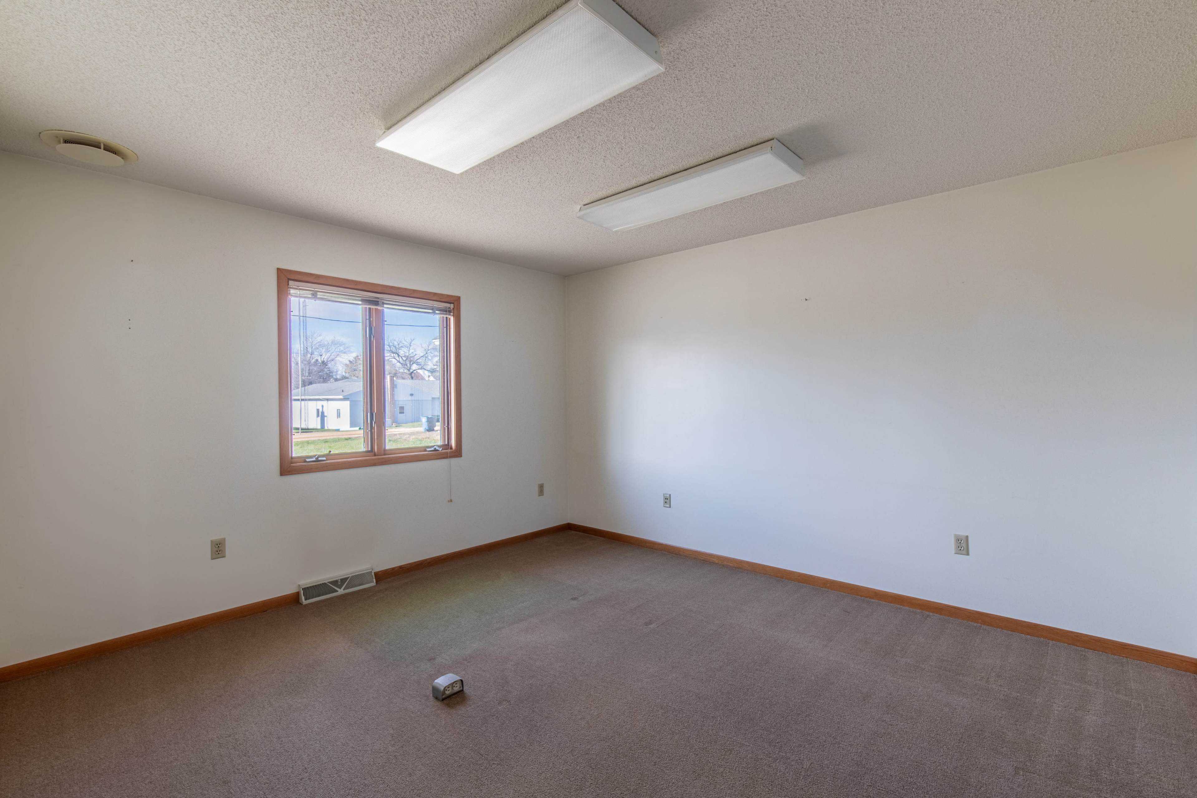 An empty room with beige carpet, white walls, and a single window allows natural light to enter.