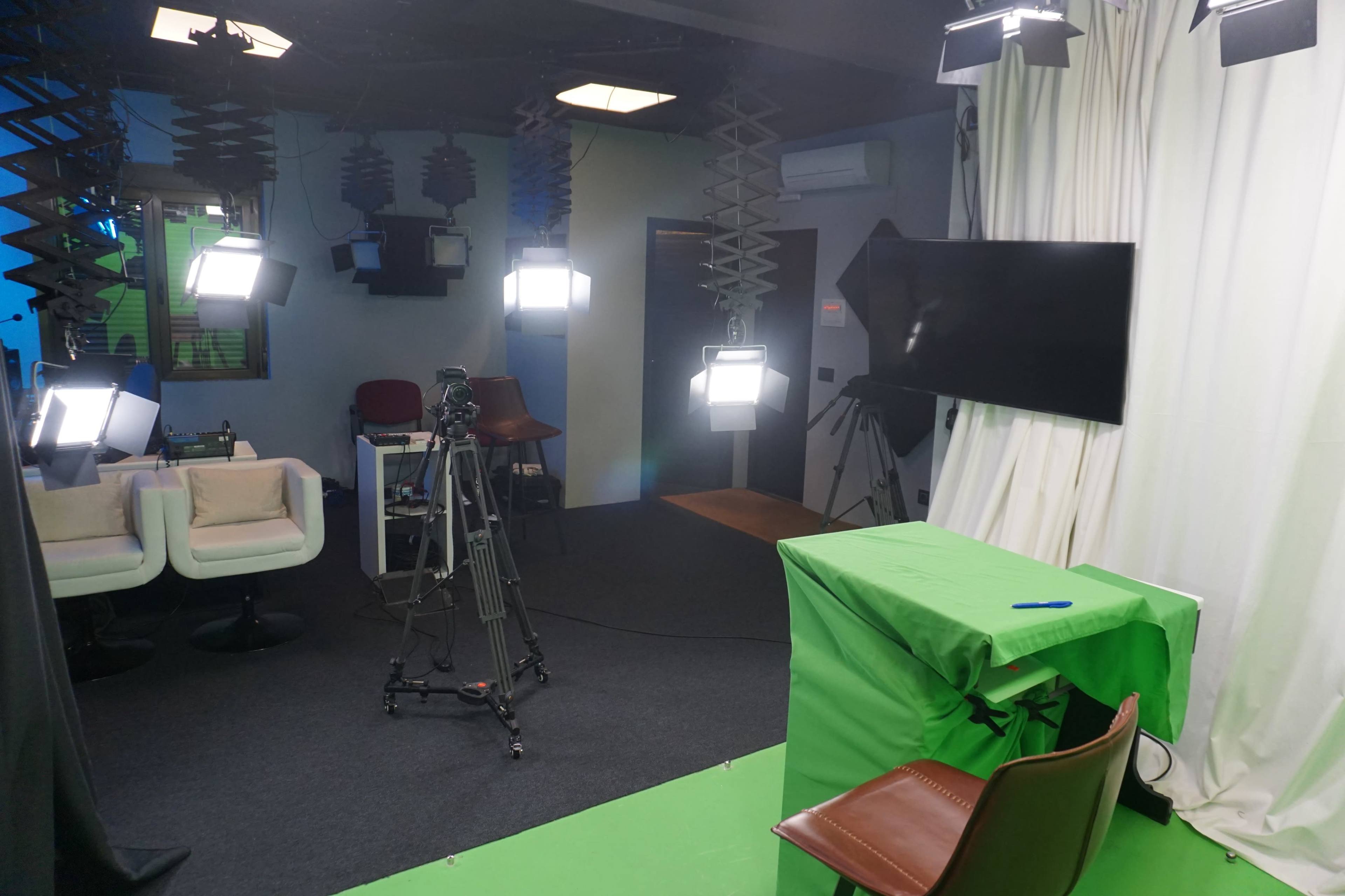 The image shows a studio setup with bright lights, a green table covered by a cloth, a camera on a tripod, and a blank screen in a room designed for video production.