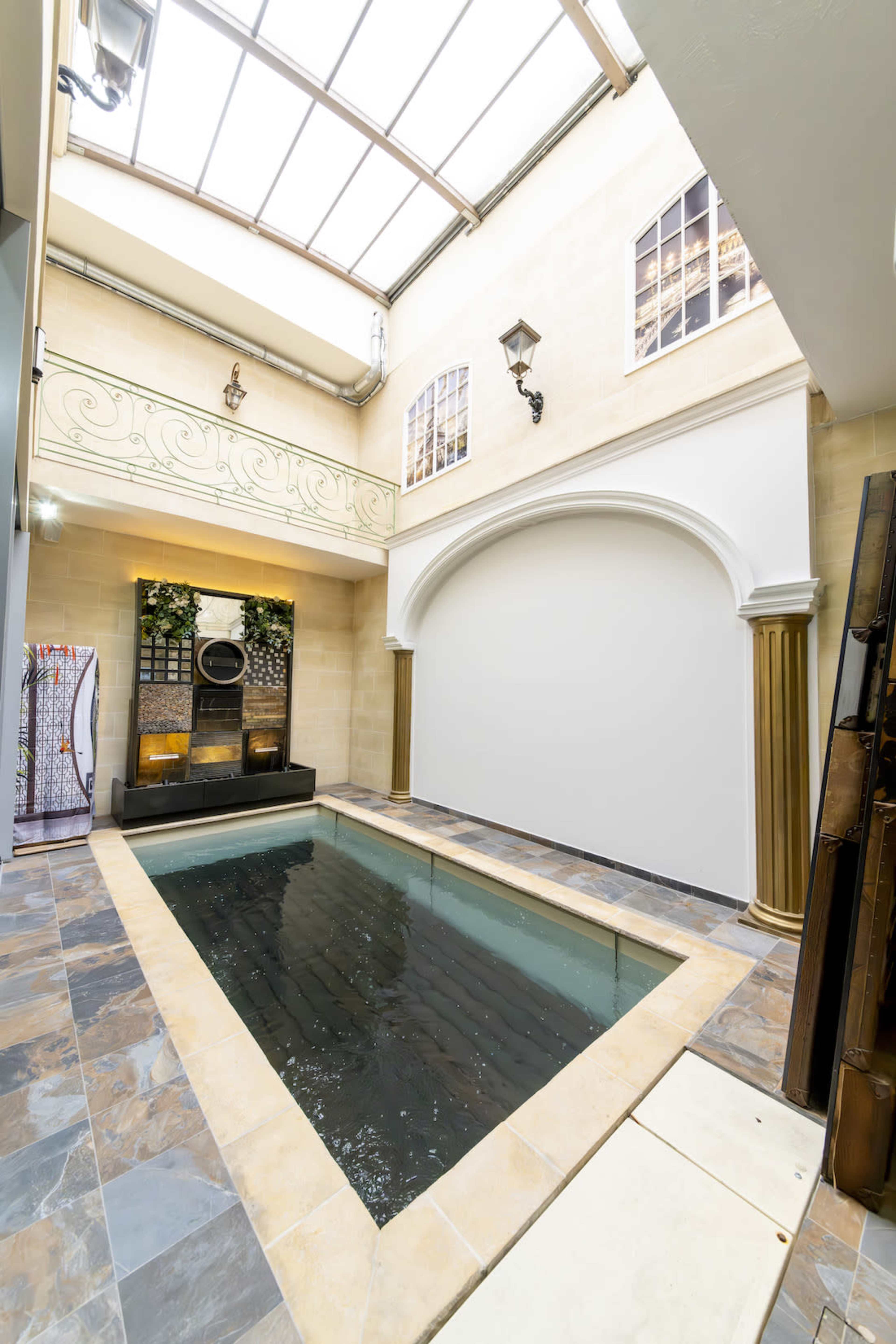 The image shows an indoor swimming pool bordered by stone walls and adorned with columns and large windows overhead.